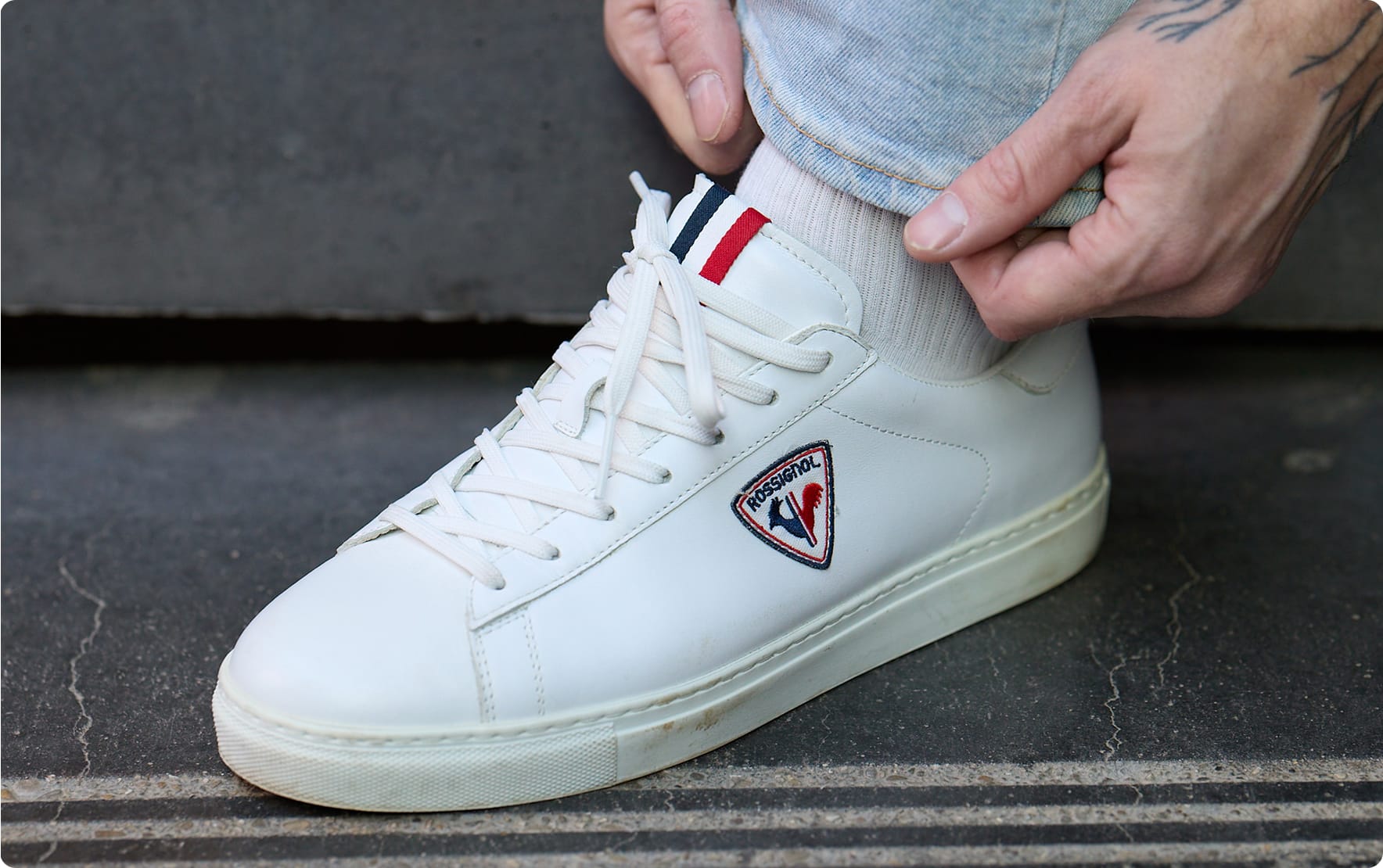 Gros plan sur une sneaker blanche portée au pied, avec lacets blancs et logo latéral, posée sur un sol en béton.