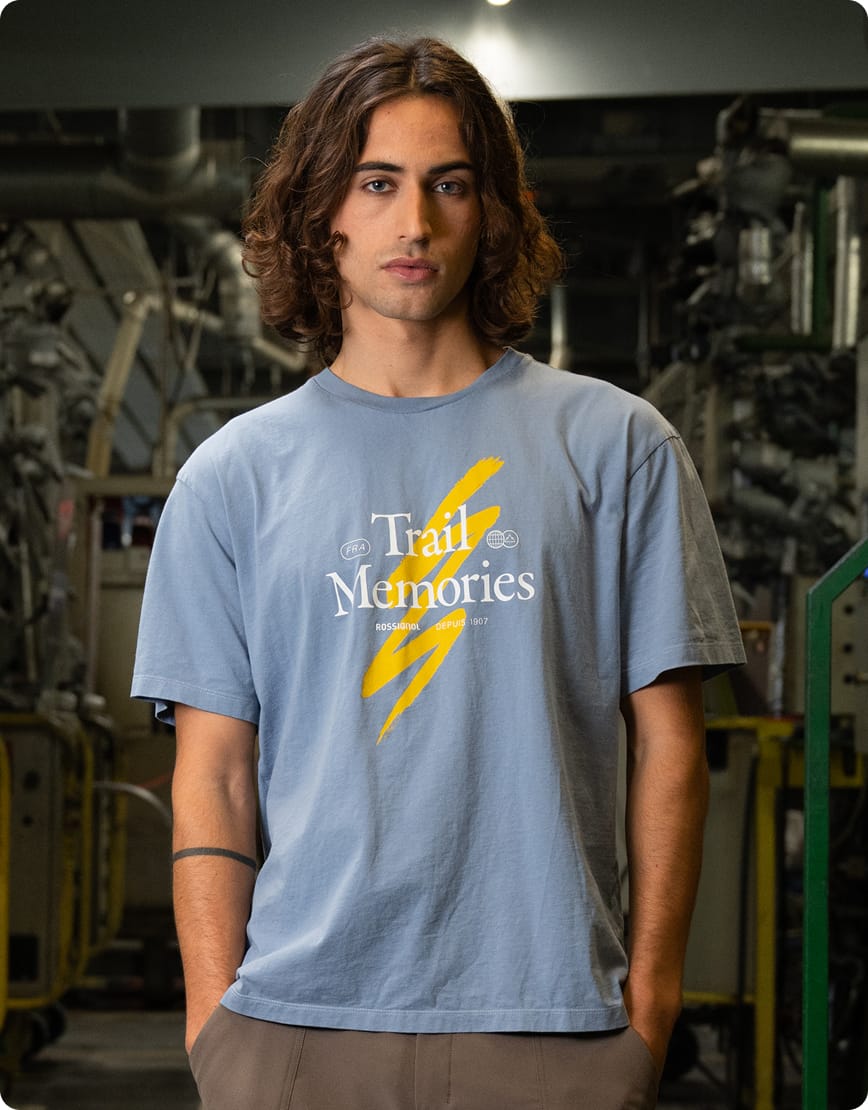 Homme portant un t-shirt bleu à manches courtes avec un motif graphique sur la poitrine, photographiée dans un atelier industriel.