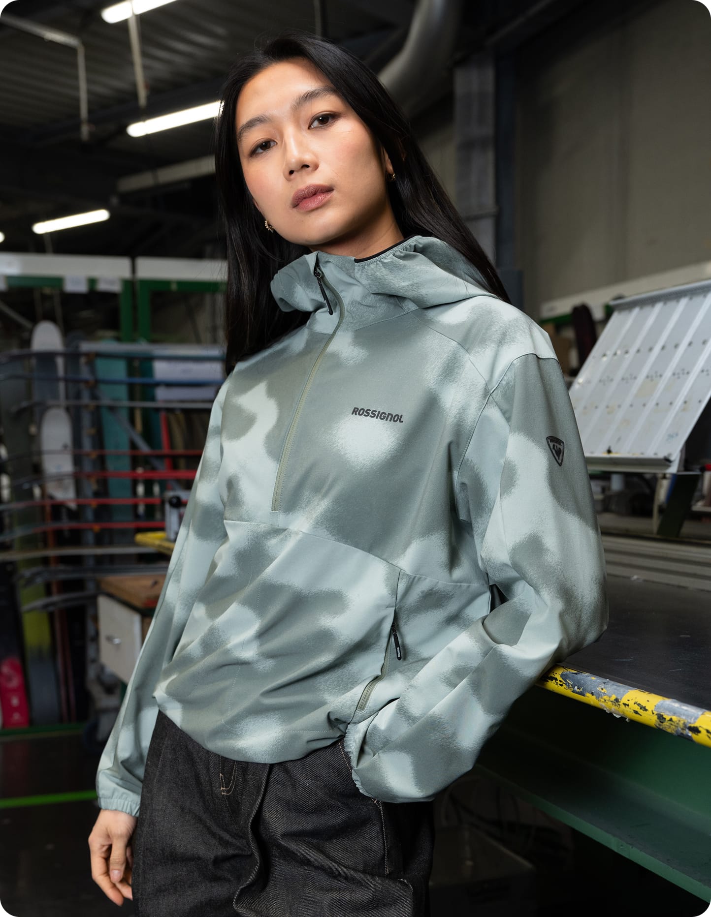 Femme portant un anorak stretch à capuche gris clair, debout dans un environnement industriel avec des machines en arrière-plan.
