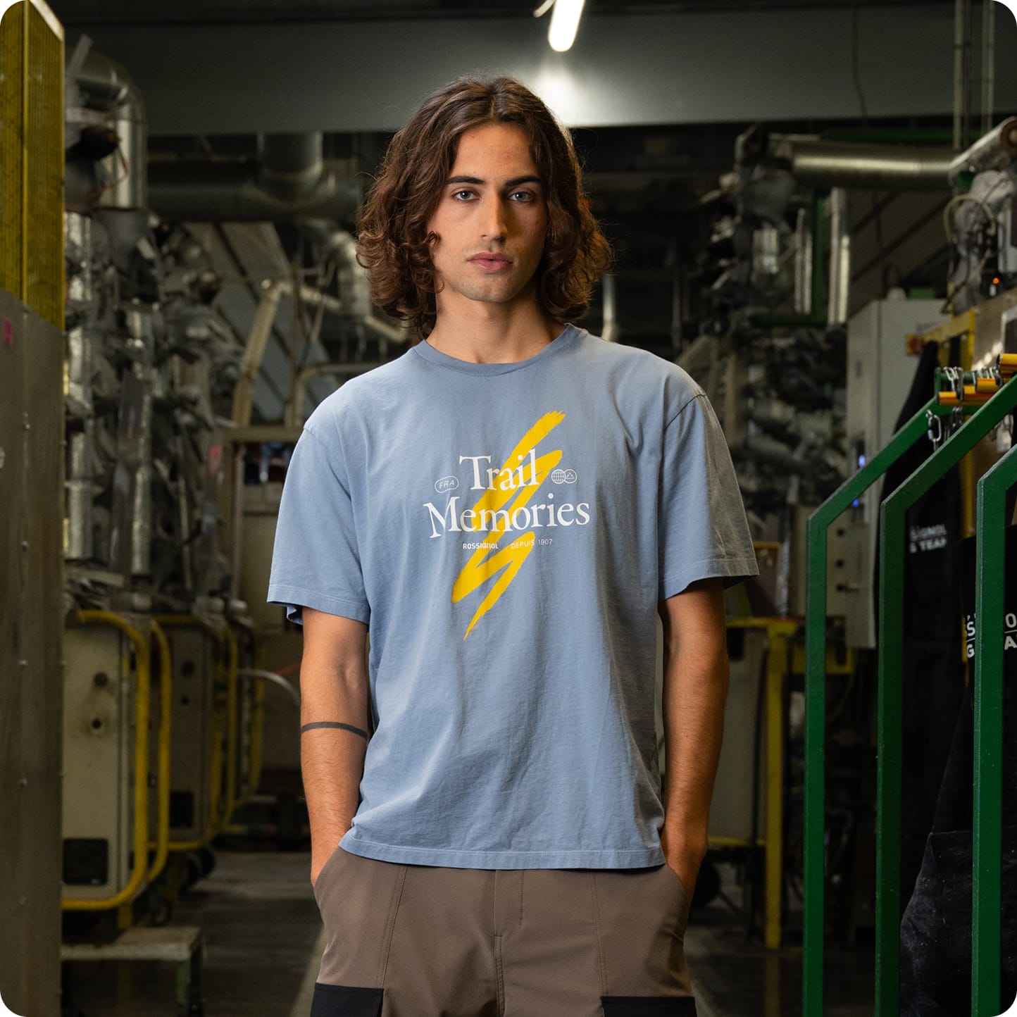 Homme portant un t-shirt bleu à manches courtes avec un motif graphique sur la poitrine, photographiée dans un atelier industriel.