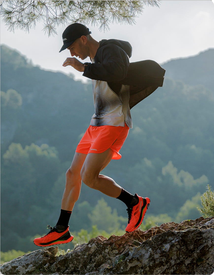 Personne en tenue de trail Rossignol avec short rouge et chaussures assorties sautant sur un rocher en pleine nature, avec des collines verdoyantes en arrière-plan.