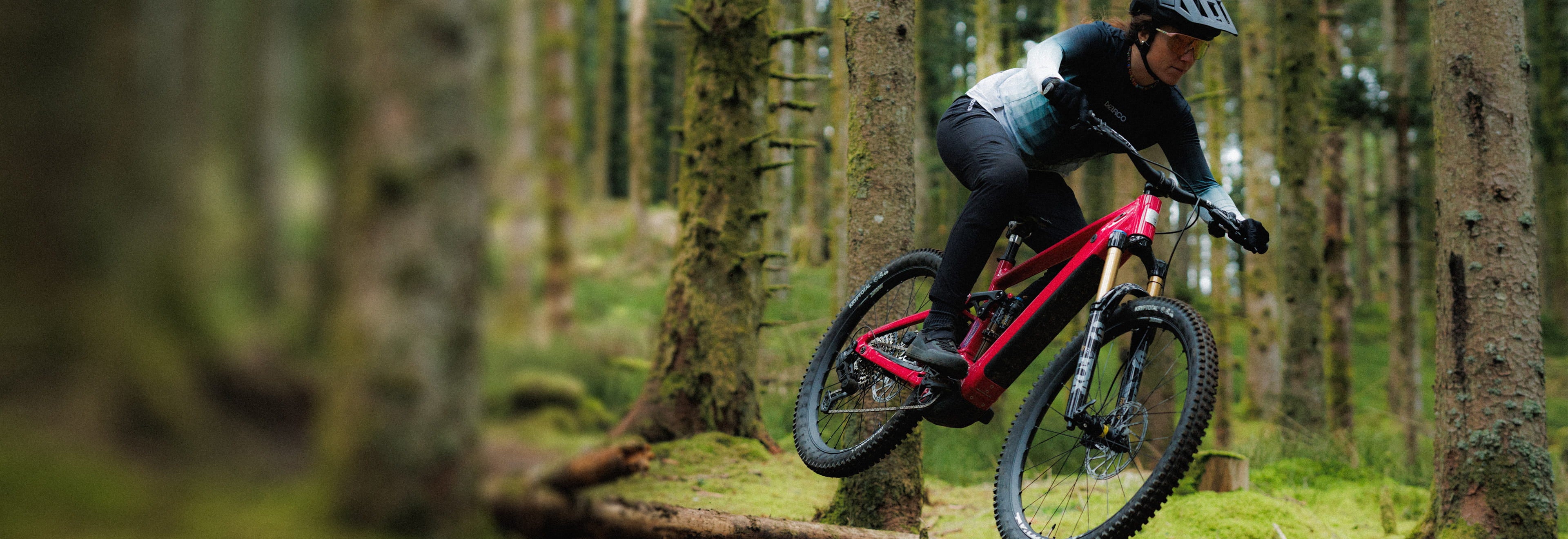 A Rossignol athlete with her After Hours mountain bike riding over a large root in the middle of a lush green forest.