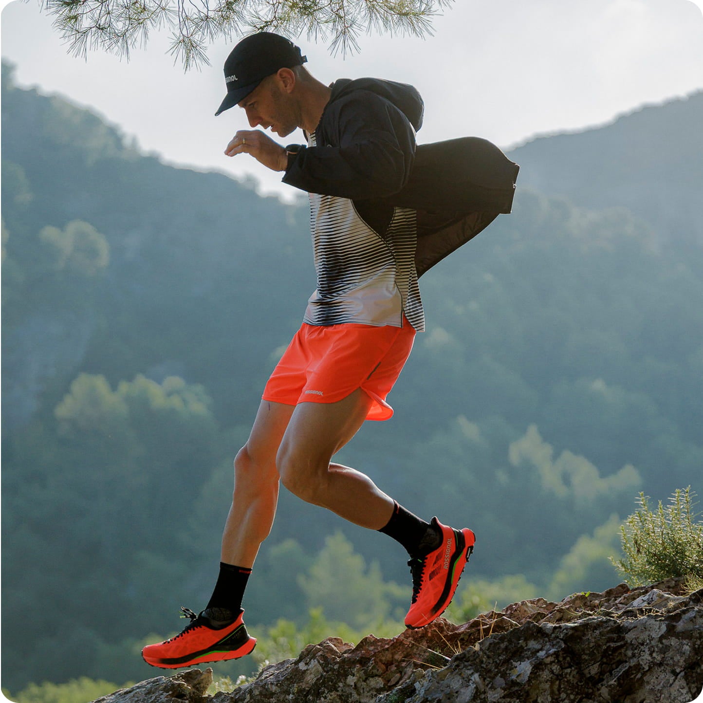 Personne en tenue de trail Rossignol avec short rouge et chaussures assorties sautant sur un rocher en pleine nature, avec des collines verdoyantes en arrière-plan.
