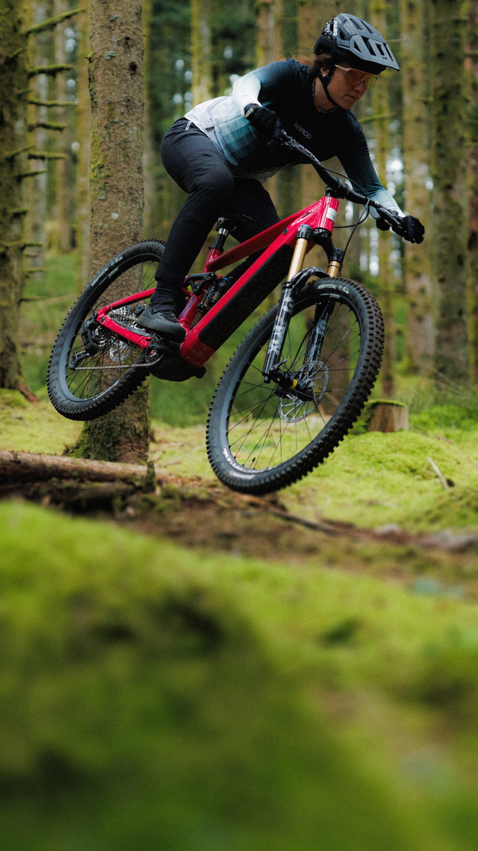 A Rossignol athlete with her After Hours mountain bike riding over a large root in the middle of a lush green forest.