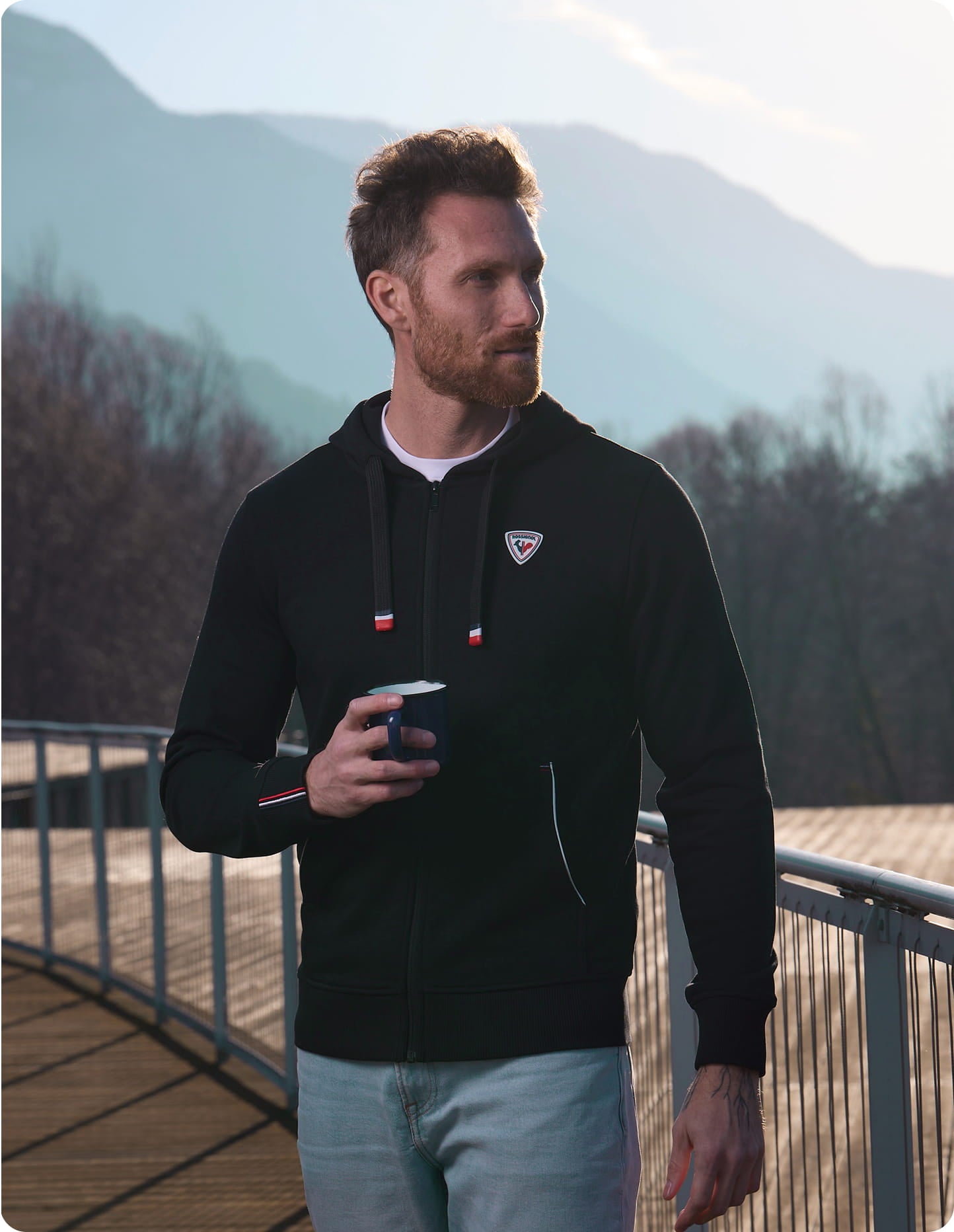 Personne portant une veste noire Rossignol et un pantalon clair, tenant un petit objet, marchant sur une terrasse en bois avec des montagnes en arrière-plan.