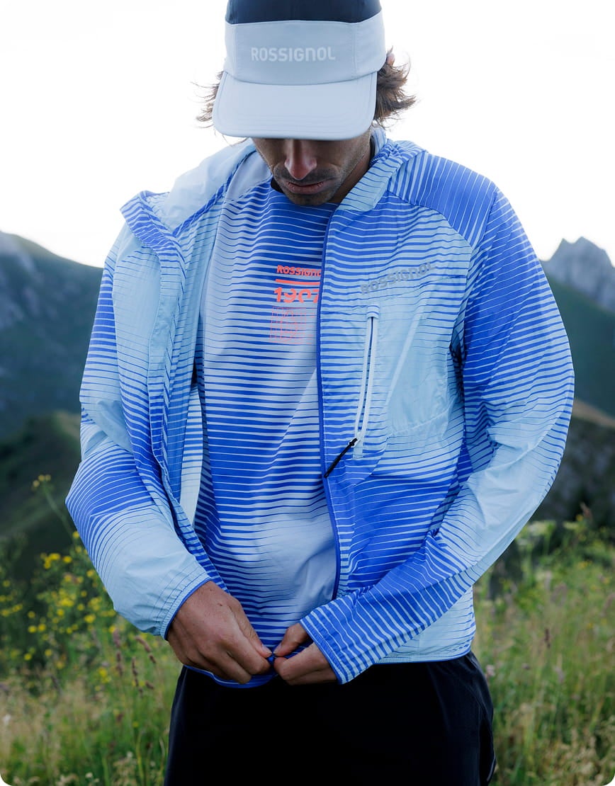 Personne portant une veste légère de trail à rayures bleues et une visière, en extérieur dans un environnement montagneux.