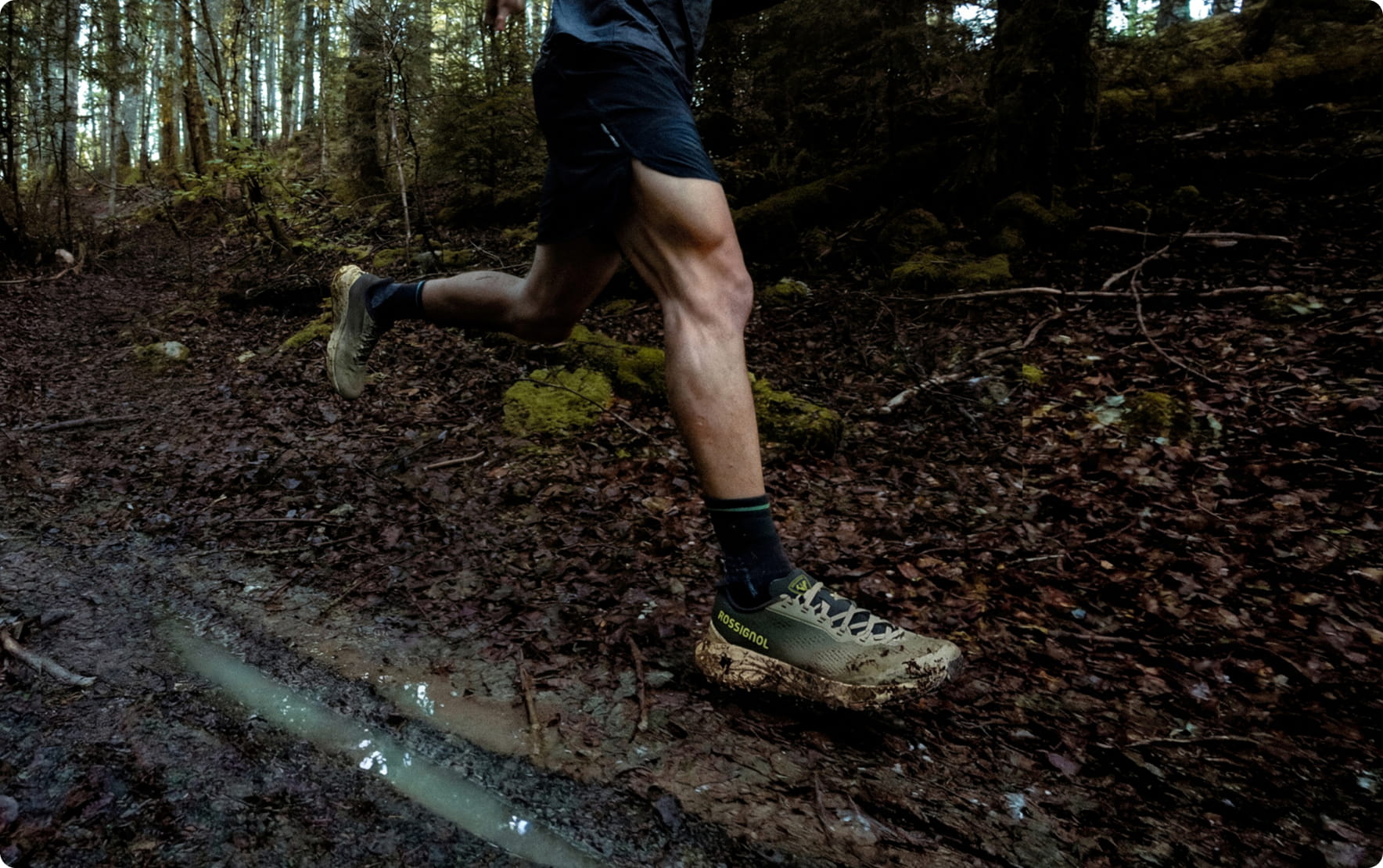 Gros plan sur les jambes d’une personne courant sur un sentier forestier humide et boueux, chaussée de chaussures de trail adaptées aux terrains difficiles.