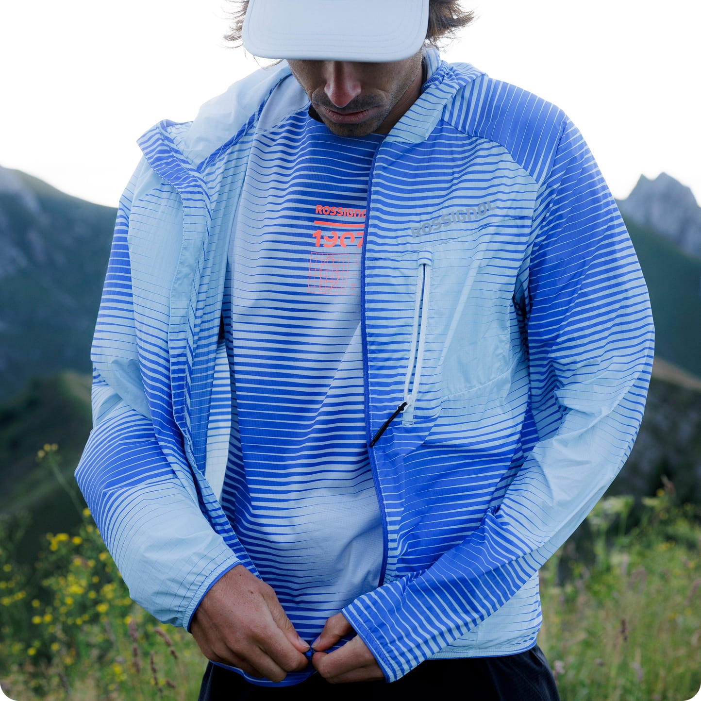 Personne portant une veste légère de trail à rayures bleues et une visière, en extérieur dans un environnement montagneux.