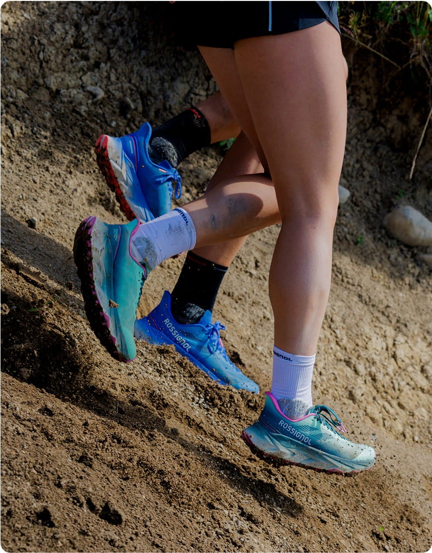 Primo piano di due persone che corrono su un sentiero sterrato indossando scarpe da trail Rossignol nei colori blu e turchese, su un terreno naturale accidentato.