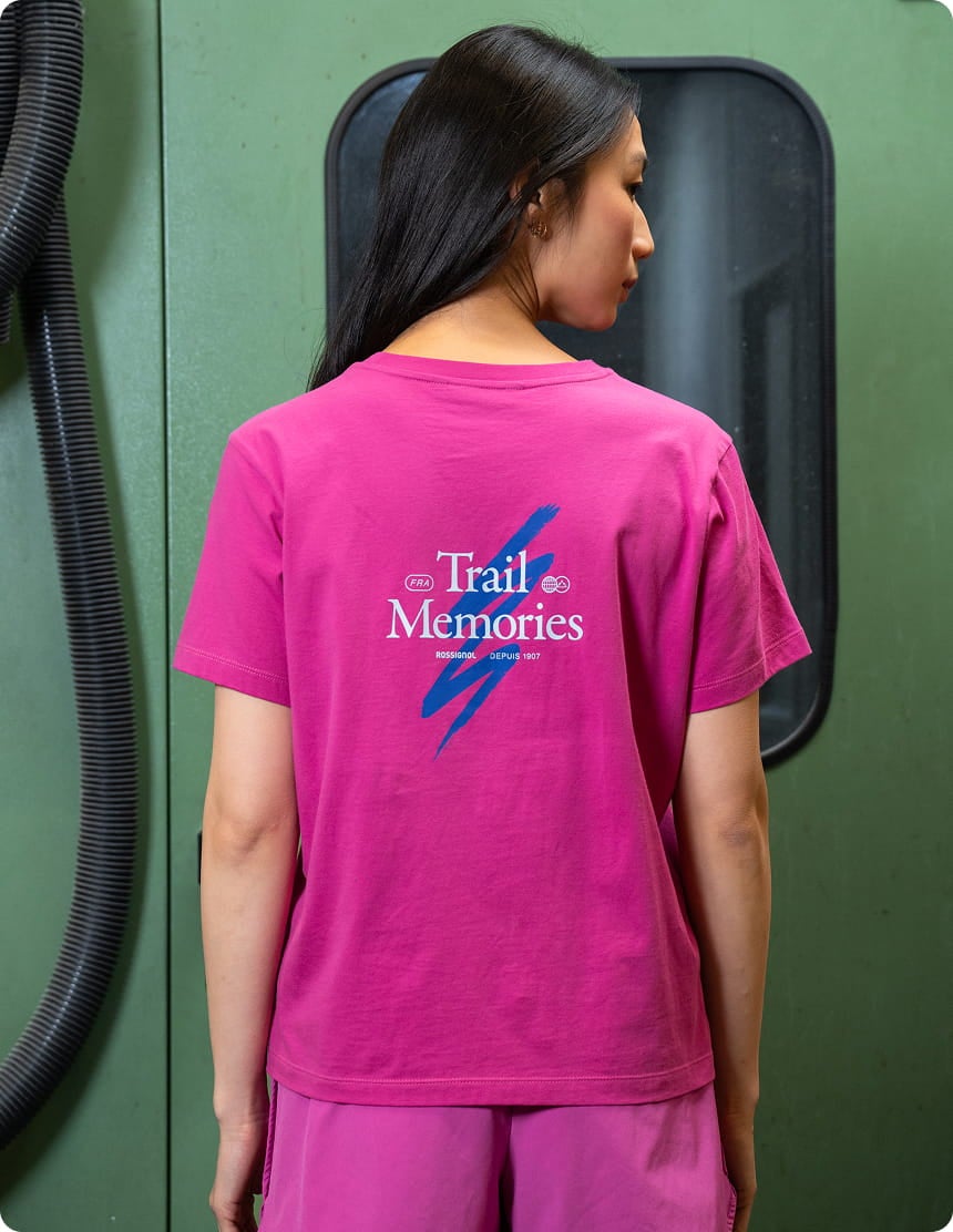 Persona che indossa una T-shirt rosa Rossignol vista da dietro, con la grafica “Trail Memories” stampata al centro della schiena. La foto è stata scattata in un contesto industriale.