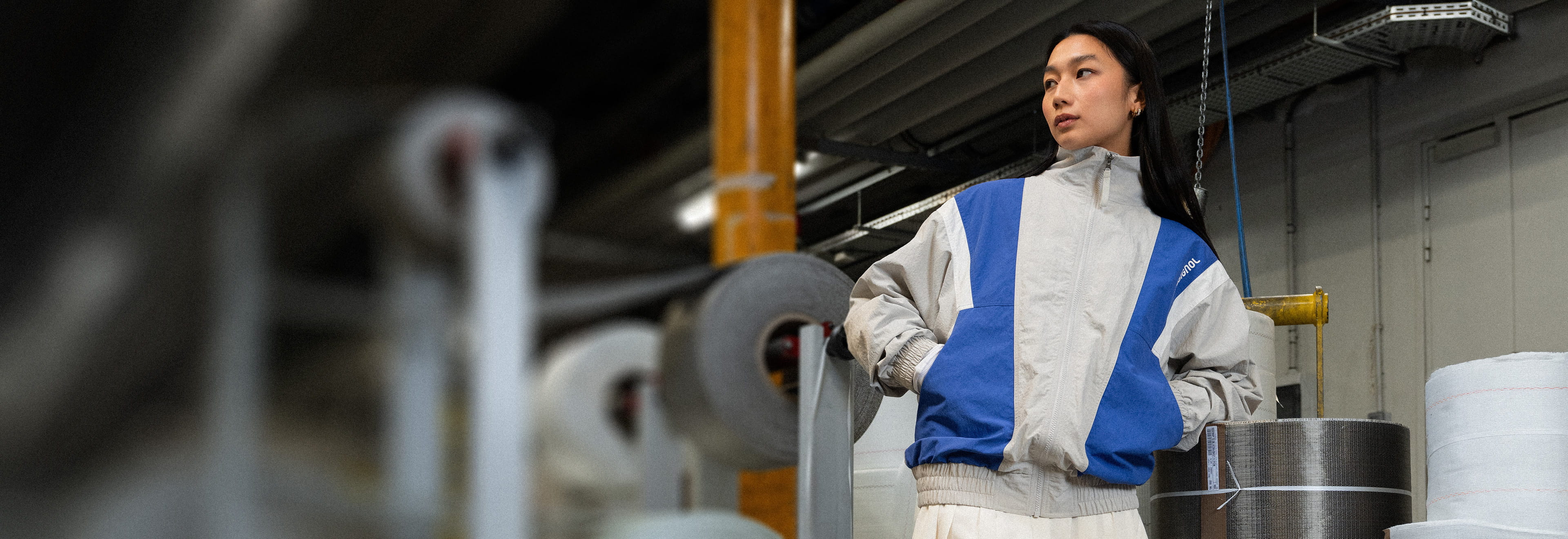 Persona giovane che indossa una giacca rétro Rossignol con zip nei colori grigio e blu, caratterizzata da una vestibilità ampia ispirata all'abbigliamento da sci vintage. La giacca è fotografata in un contesto industriale.