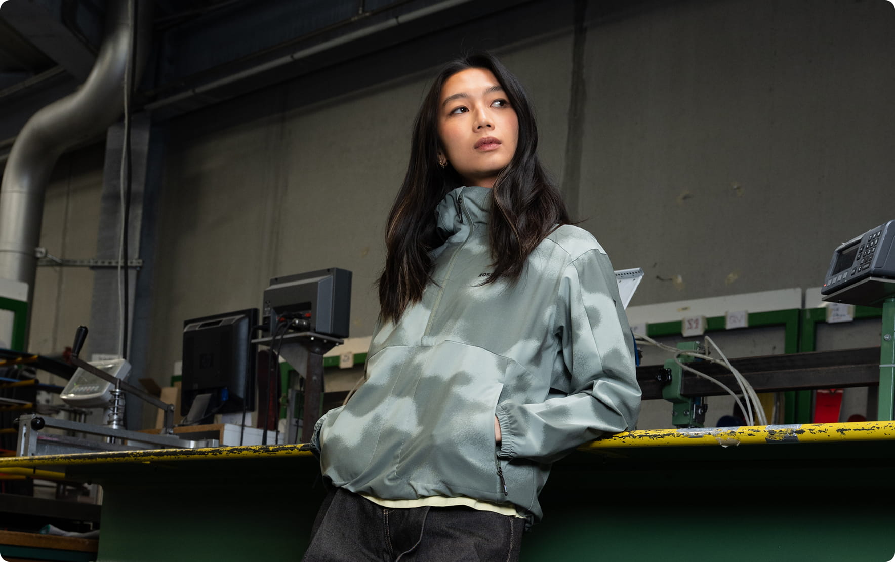 Person wearing a light green hooded anorak, leaning against a counter in an industrial environment. Technical equipment is visible in the background within a production workshop.