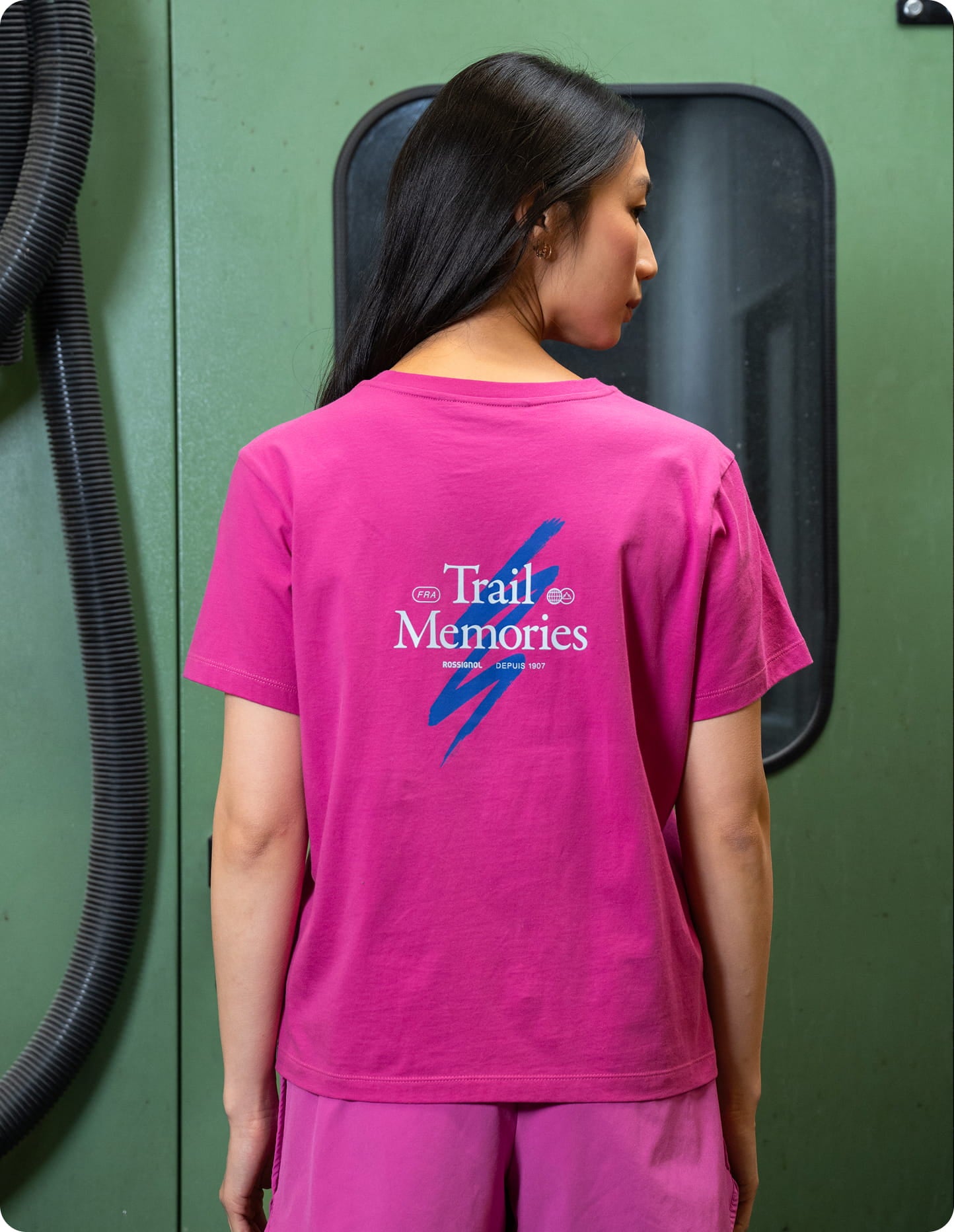Persona che indossa una T-shirt rosa Rossignol vista da dietro, con la grafica “Trail Memories” stampata al centro della schiena. La foto è stata scattata in un contesto industriale.