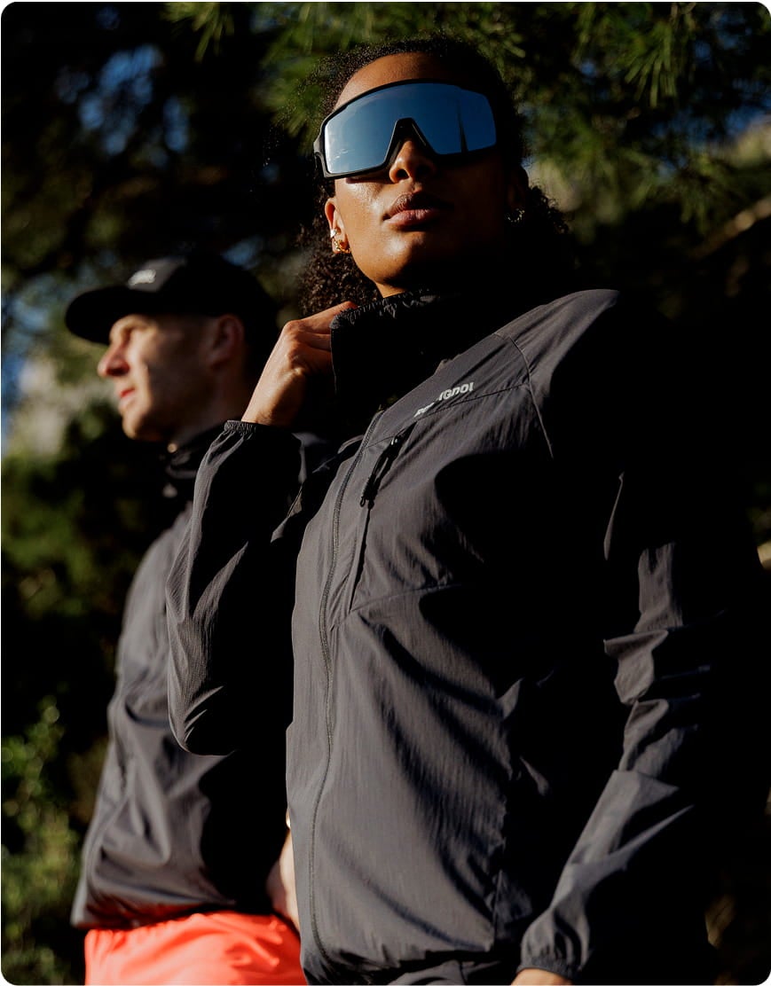 Deux personnes portant des vestes techniques de trail en extérieur, dont l'une avec des lunettes de sport à écran, dans un environnement montagneux.