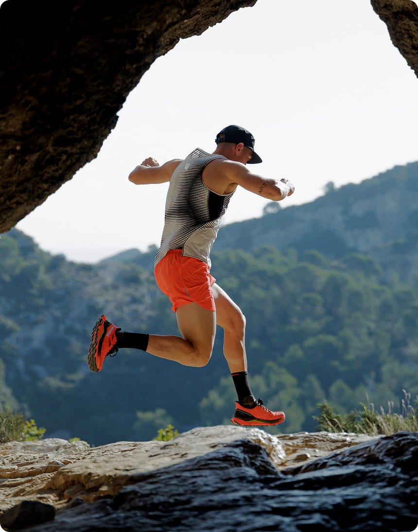 Coureur en plein saut sur un sentier rocheux en montagne, portant une tenue de trail légère avec un paysage naturel en arrière-plan.