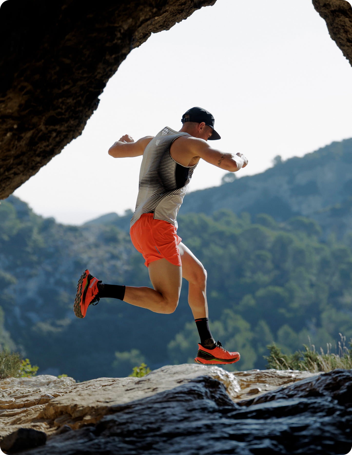 Coureur en plein saut sur un sentier rocheux en montagne, portant une tenue de trail légère avec un paysage naturel en arrière-plan.