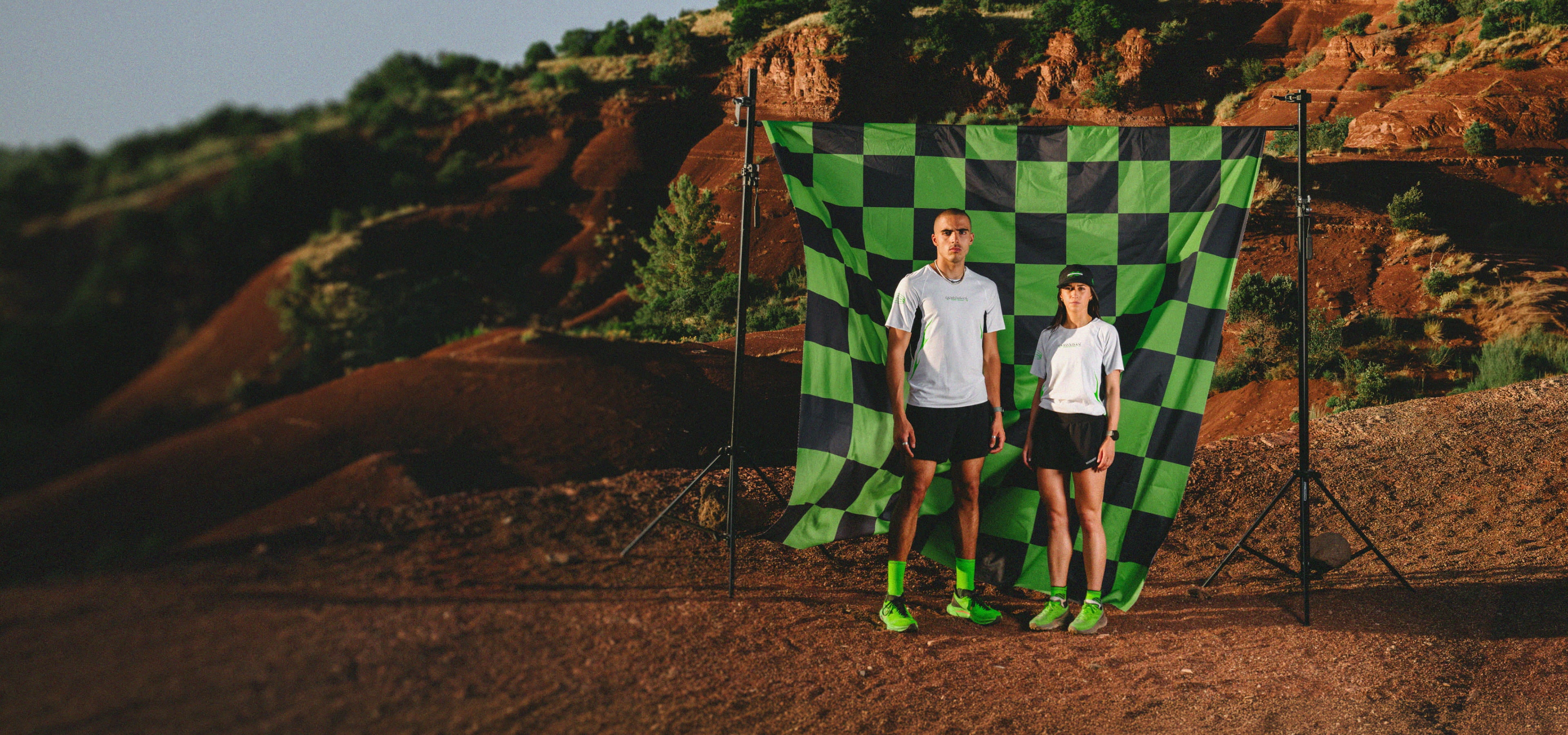 Two people standing in front of a large green and black checkered flag, resembling a race flag. They are wearing Rossignol outfits from the Super Project trail capsule.