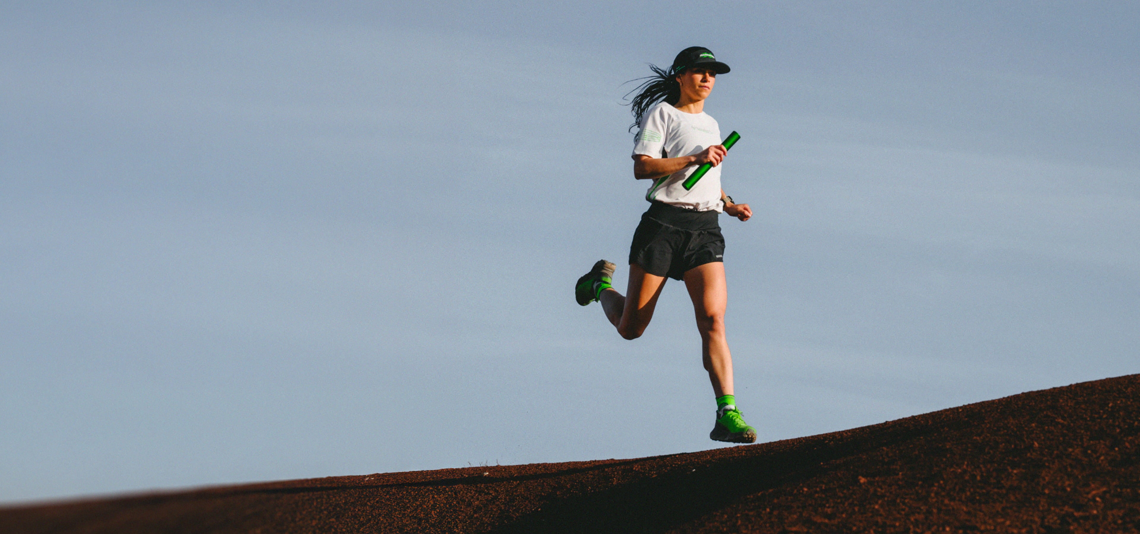 A woman trail running holding a relay baton and wearing the limited edition "Super Project" Rossignol Vercors shoes.