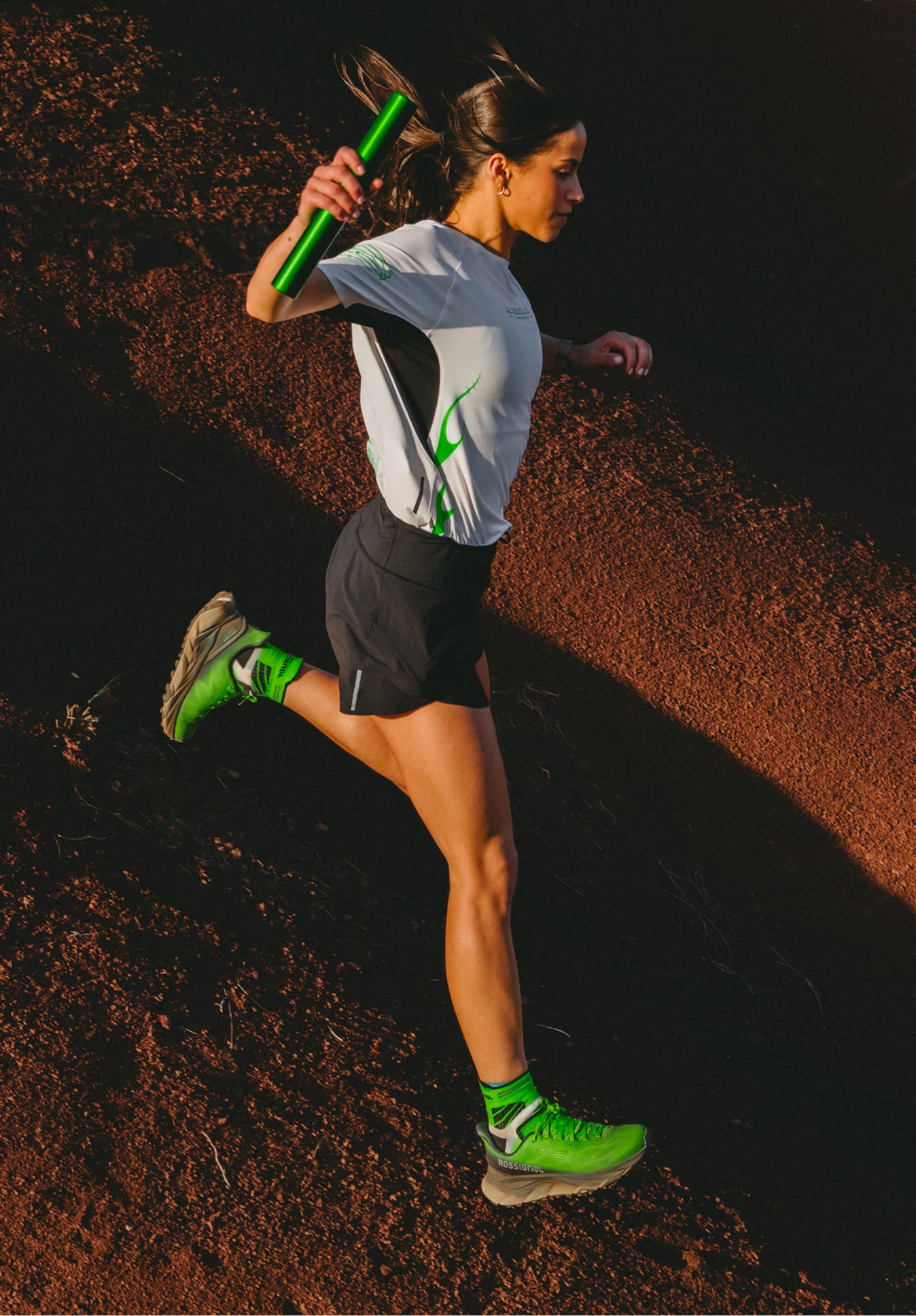 Athlete wearing the Rossignol SAPA ULTRA TEE and carrying a green relay baton while running downhill on a dirt trail
