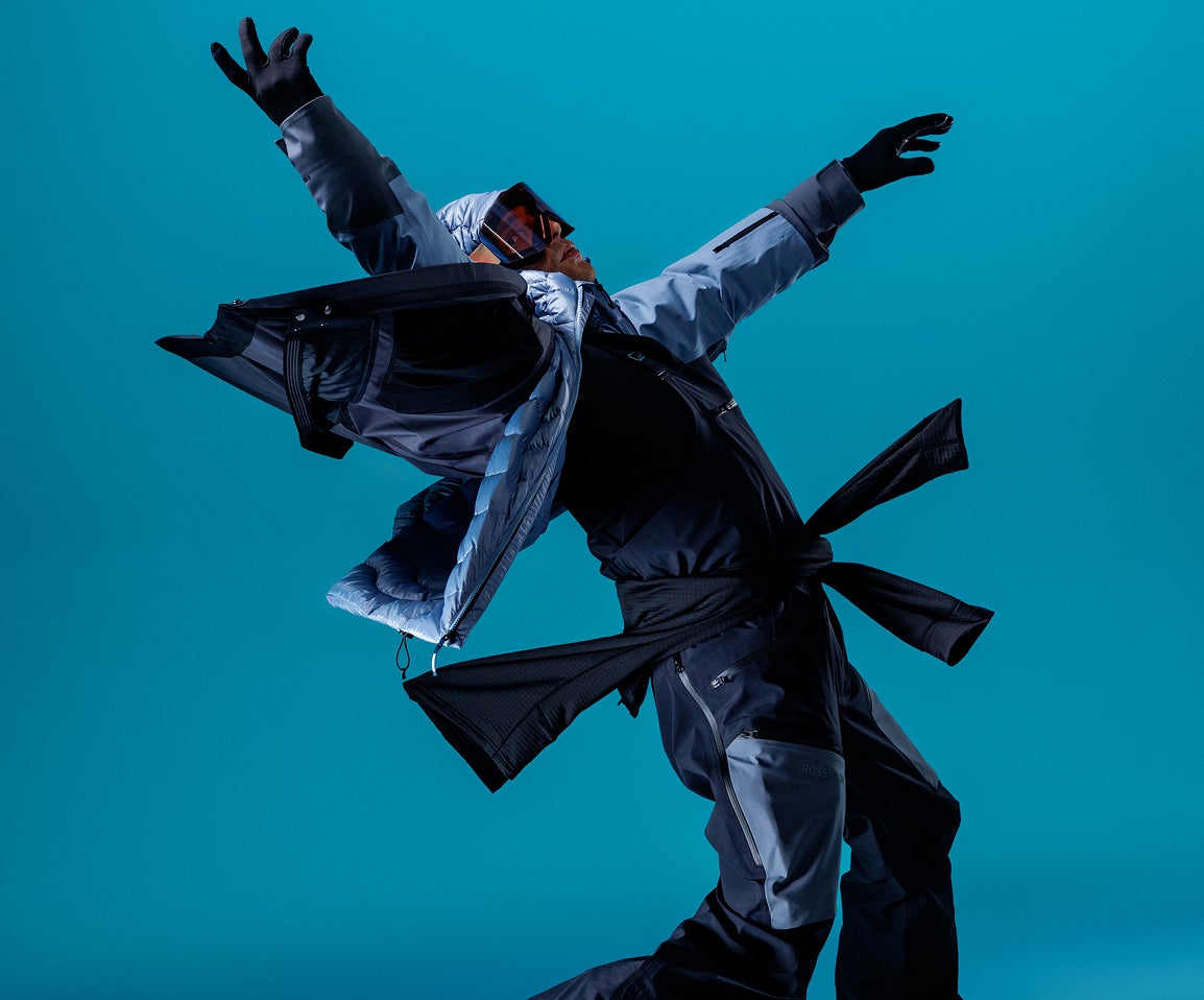 Person in Rossignol Skibekleidung in einer dynamischen Pose vor blauem Studiohintergrund, die technische Outerwear mit mehrlagiger Konstruktion präsentiert.