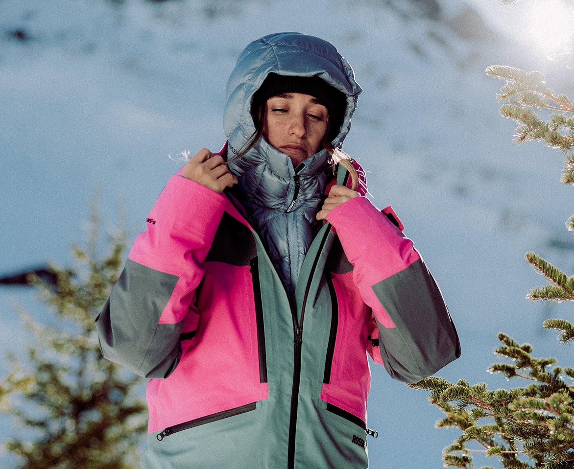 Person in einer isolierten Rossignol Skijacke, die die Kapuze in einer verschneiten Berglandschaft ajustiert.
