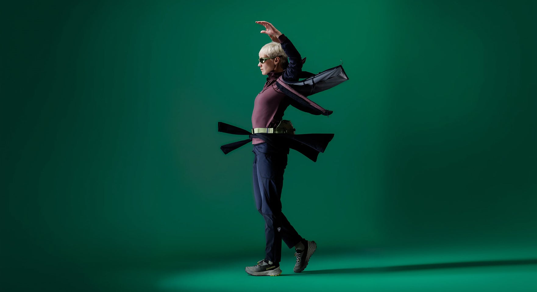 Person in Rossignol Performance-Bekleidung in einer dynamischen Pose vor grünem Studiohintergrund, die leichte Outerwear und Bewegungsfreiheit präsentiert.