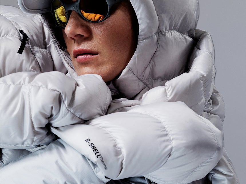 Nahaufnahme einer Person mit weißer Rossignol Daunenjacke und Performance-Brille vor einem Studiohintergrund.