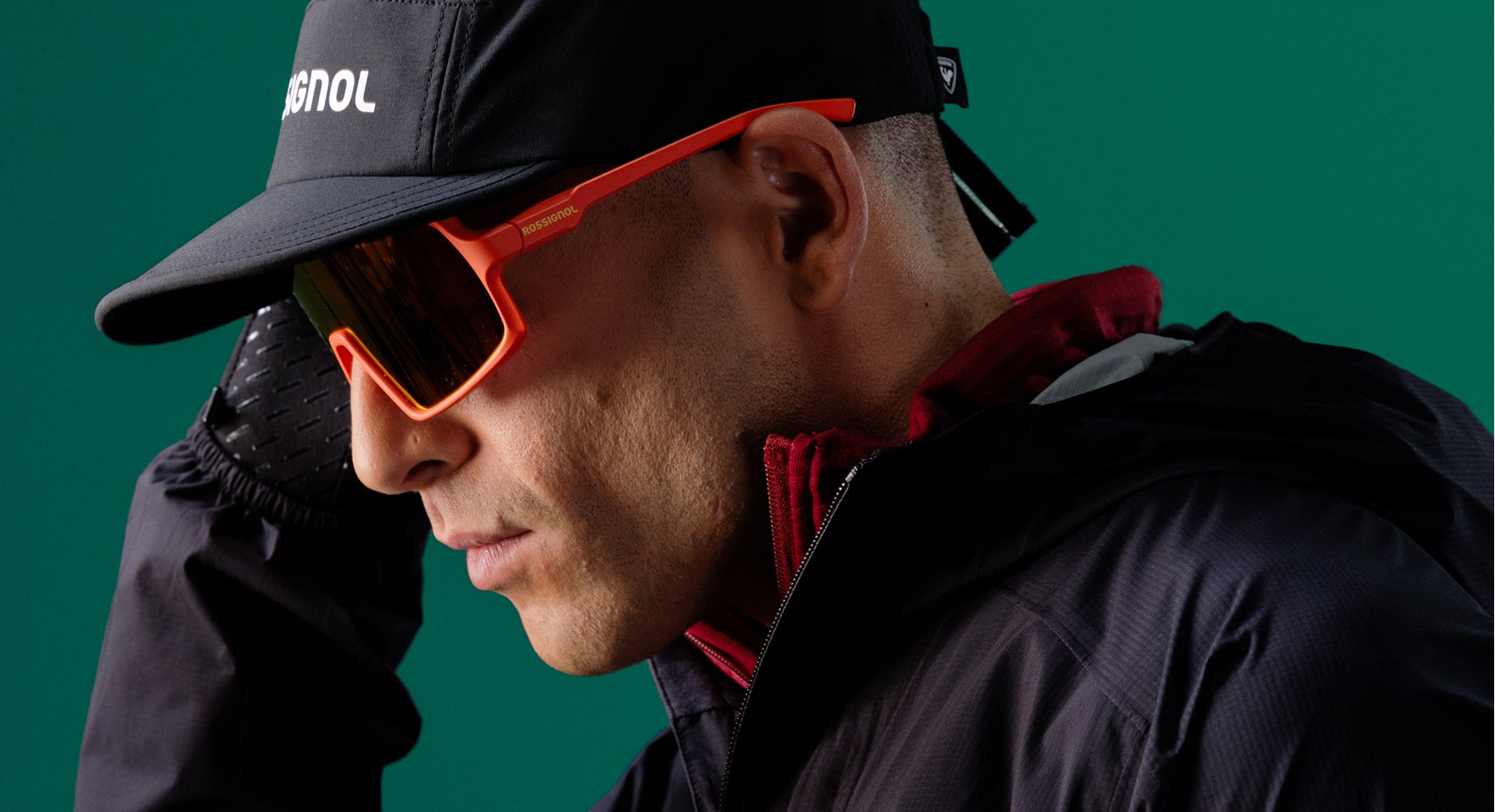 Nahaufnahme einer Person mit Rossignol Performance-Bekleidung, Kappe und Sportsonnenbrille vor grünem Studiohintergrund.