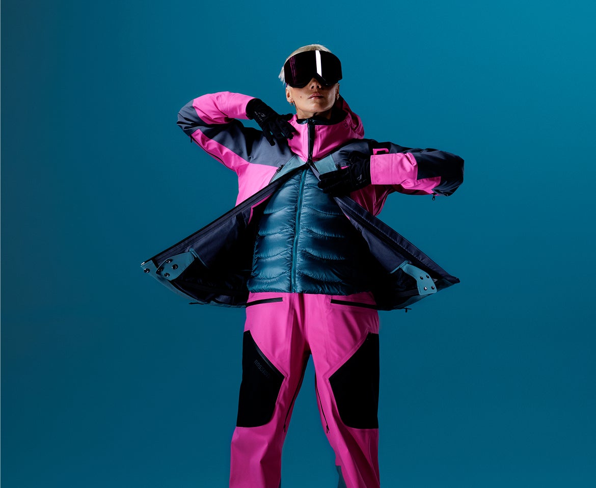 Person in einer rosa Rossignol Skibekleidung mit isolierender Zwischenschicht vor blauem Studiohintergrund.