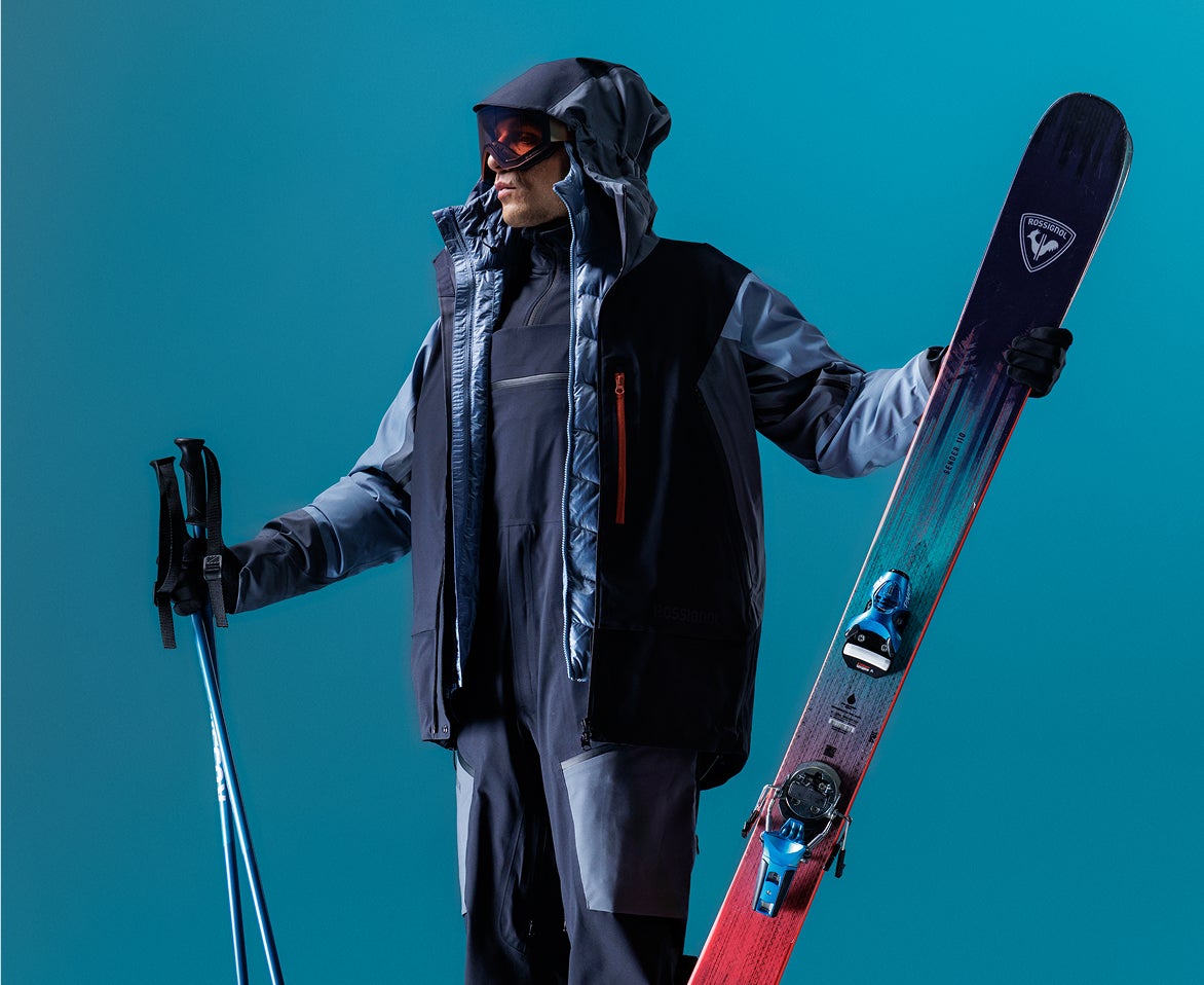 Person in Rossignol Skibekleidung mit Skiern und Skistöcken vor blauem Studiohintergrund.