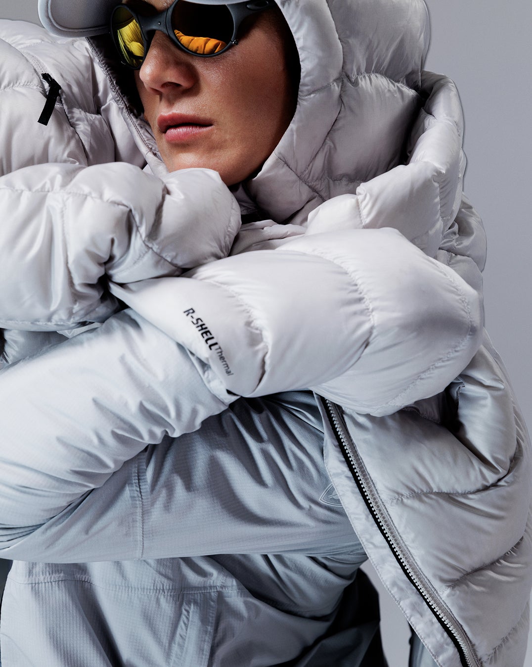 Nahaufnahme einer Person mit weißer Rossignol Daunenjacke und Performance-Brille vor einem Studiohintergrund.