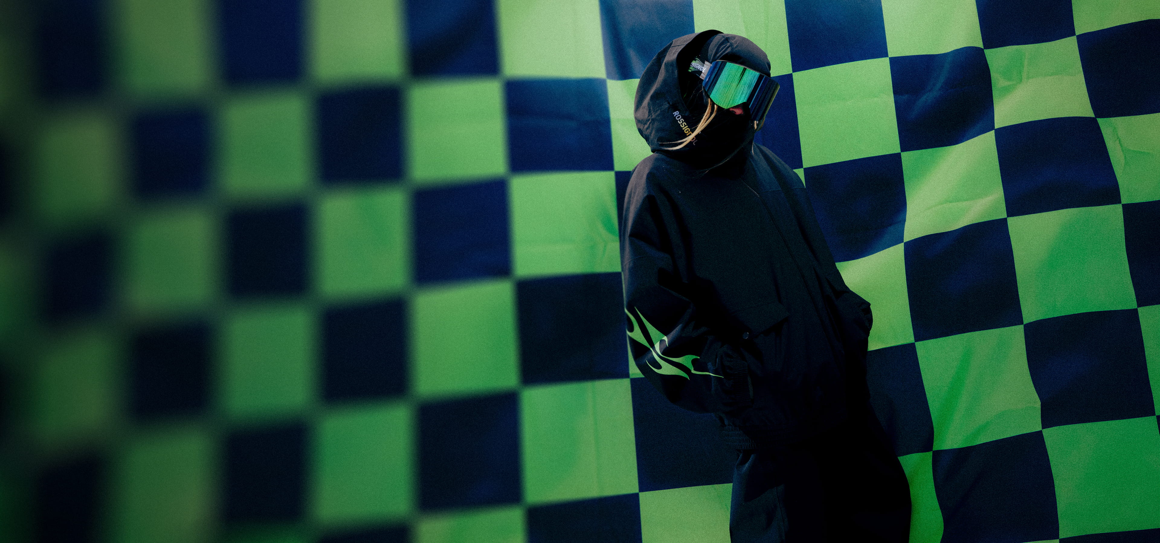 Person wearing a black ski outfit with goggles and hood in front of a green and black checkered backdrop, showcasing a limited-edition winter collection.