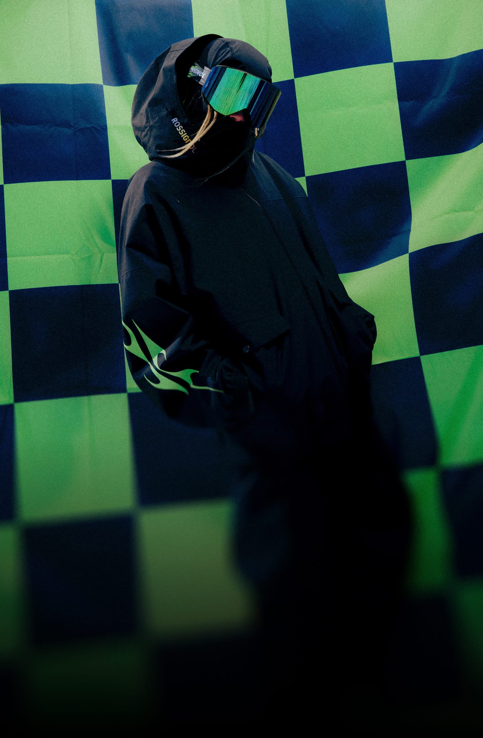 Person wearing a black ski outfit with goggles and hood in front of a green and black checkered backdrop, showcasing a limited-edition winter collection.