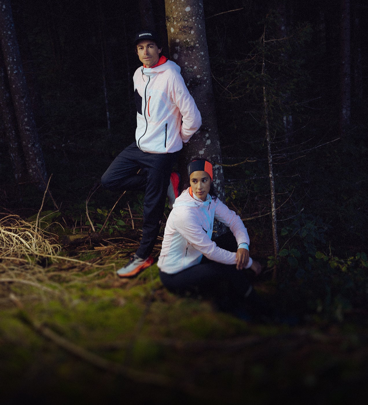 Zwei Personen tragen weiße leichte Jacken in einer Waldlandschaft. Eine Person lehnt an einem Baum, während die andere im Vordergrund sitzt.