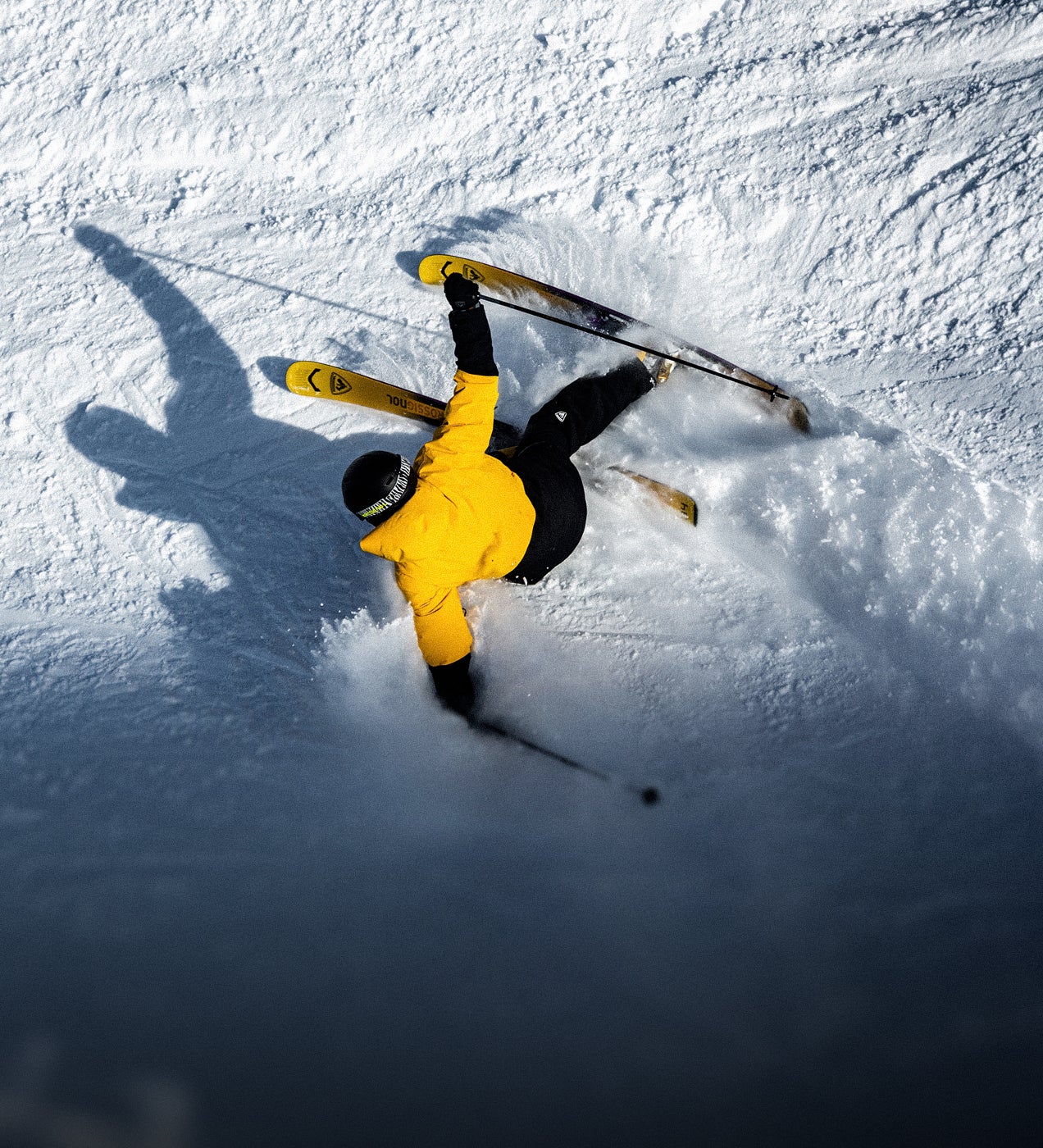Skieur vu de dessus effectuant un virage dans la neige sur une piste enneigée, portant une veste jaune.