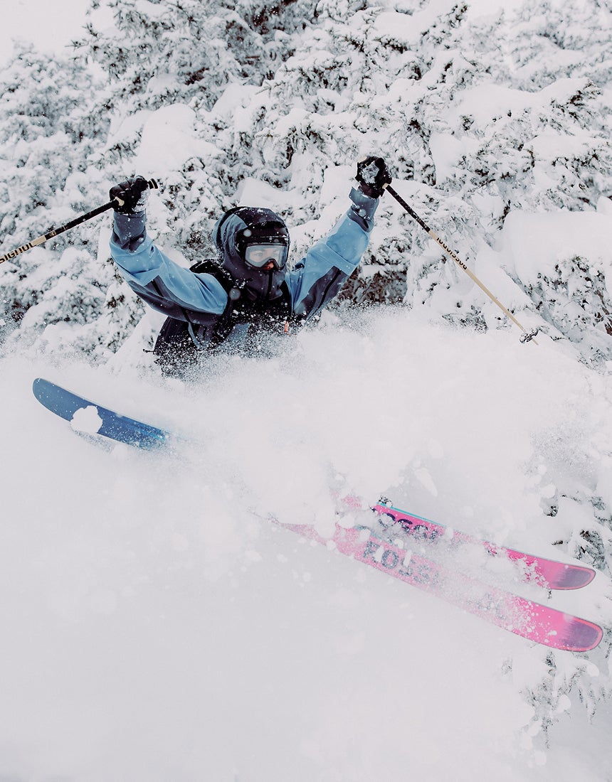A freeride skier performing a jump, kicking up a massive spray of powder, surrounded by snow-covered pine trees.