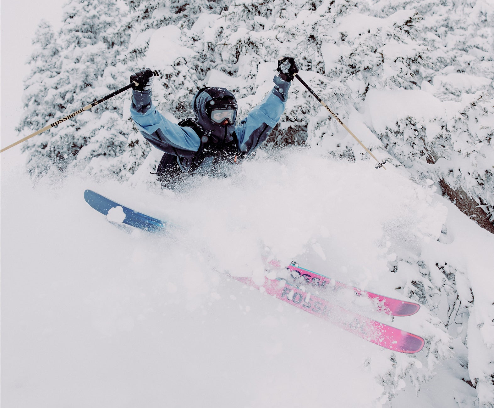 A freeride skier performing a jump, kicking up a massive spray of powder, surrounded by snow-covered pine trees.