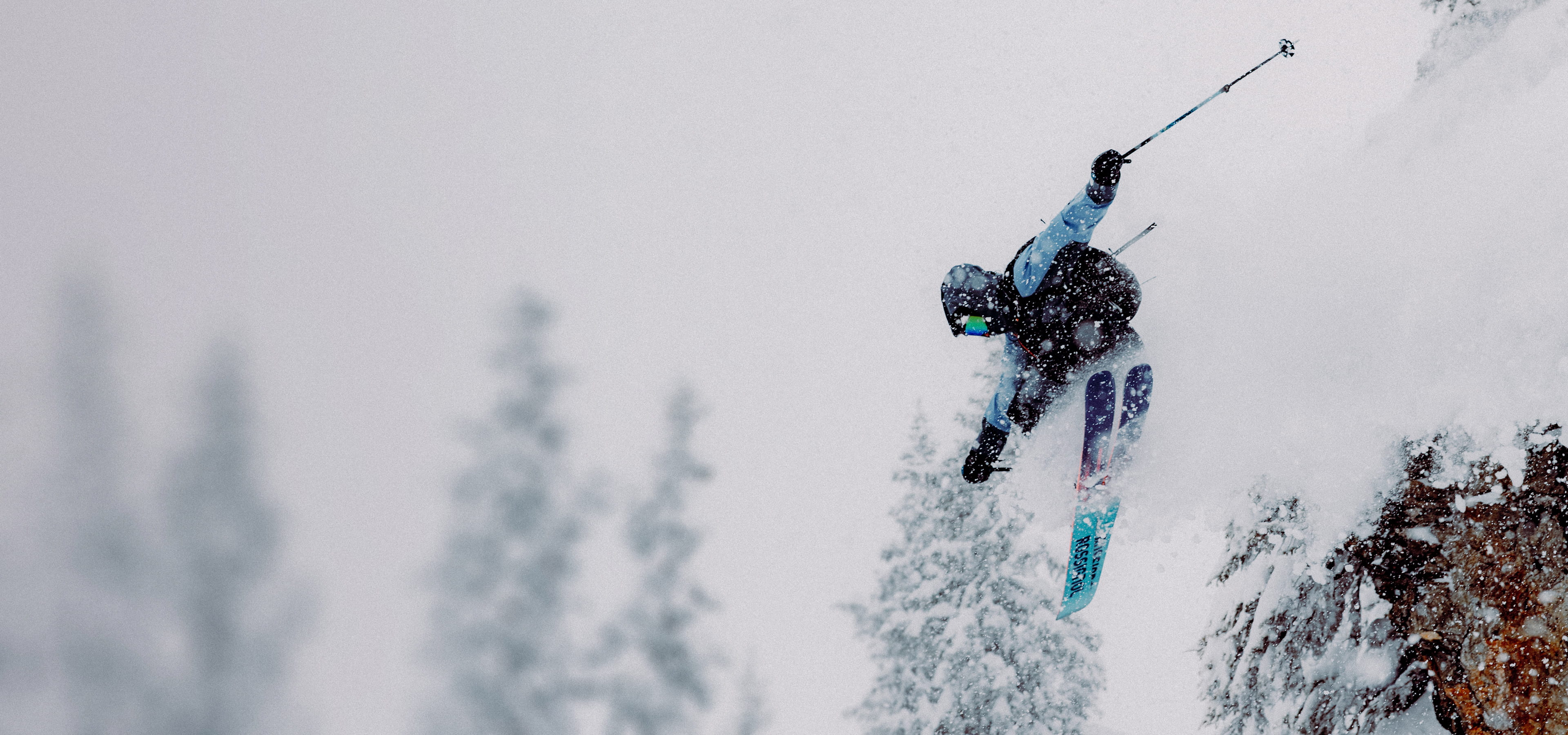 A freeride skier performing a high jump while snow falls, surrounded by snow-covered pine trees.