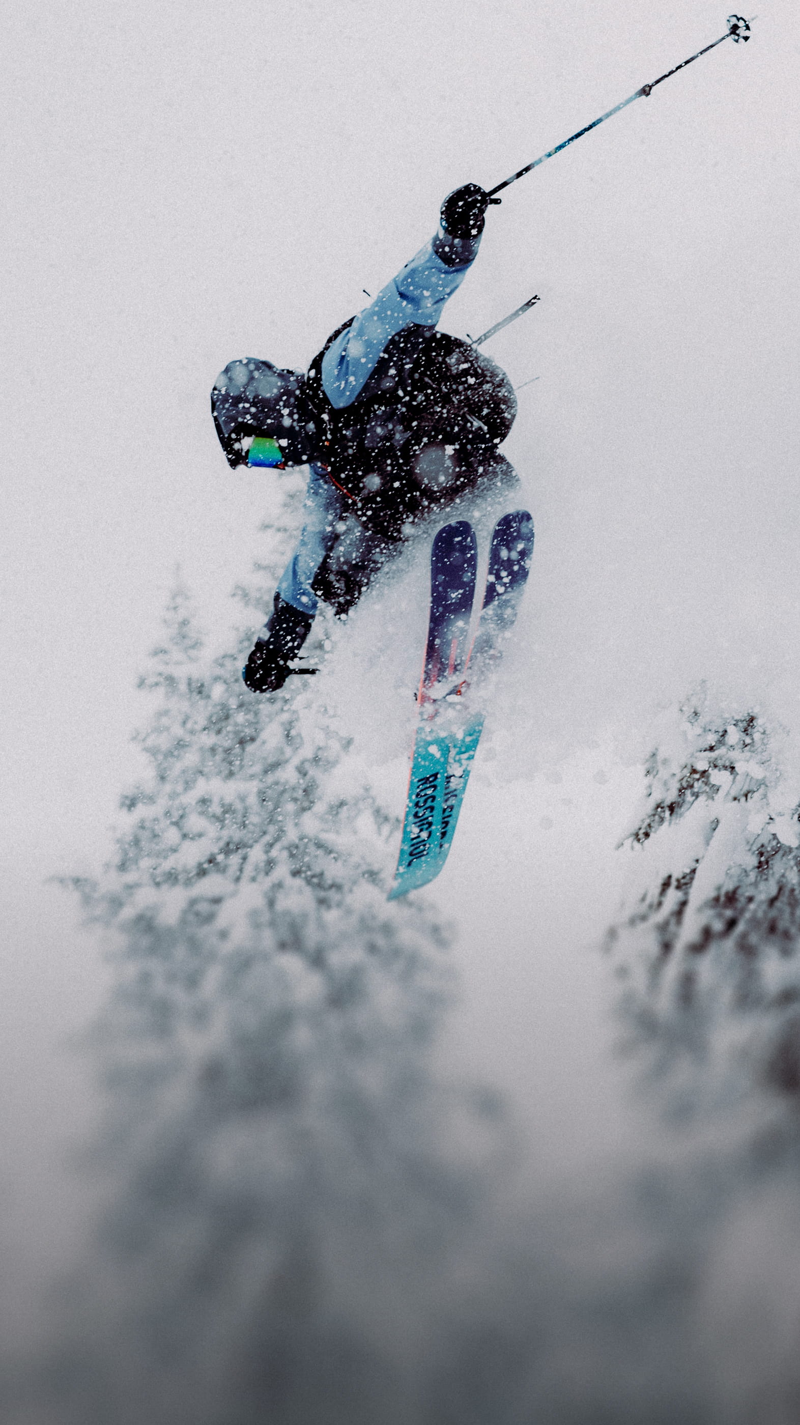 A freeride skier performing a high jump while snow falls, surrounded by snow-covered pine trees.