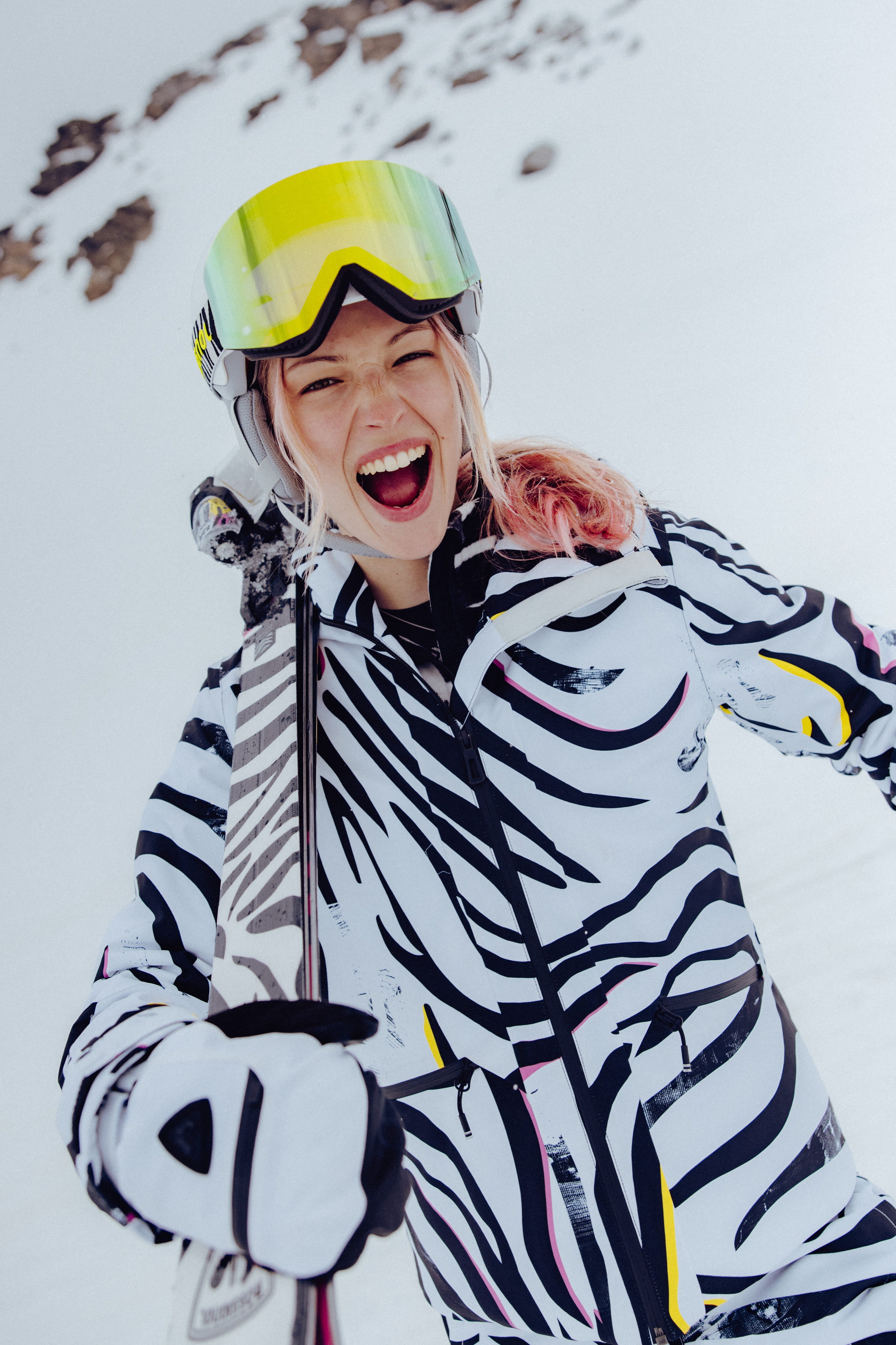 Close-up of a skier in a bold black-and-white patterned ski outfit holding a ski, wearing a white helmet and mirrored goggles on a snowy mountain slope.