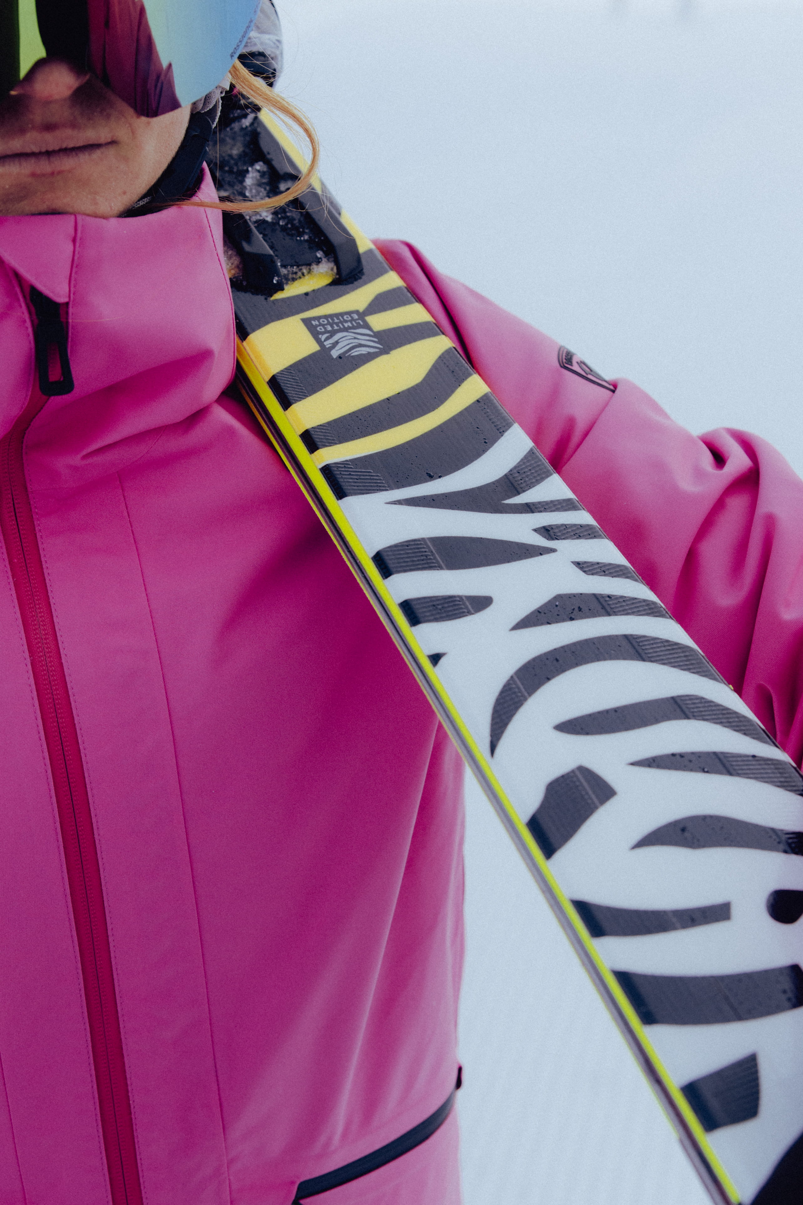 Close-up of a skier in a bright pink ski jacket carrying a ski with a bold black-and-white graphic design over her shoulder.