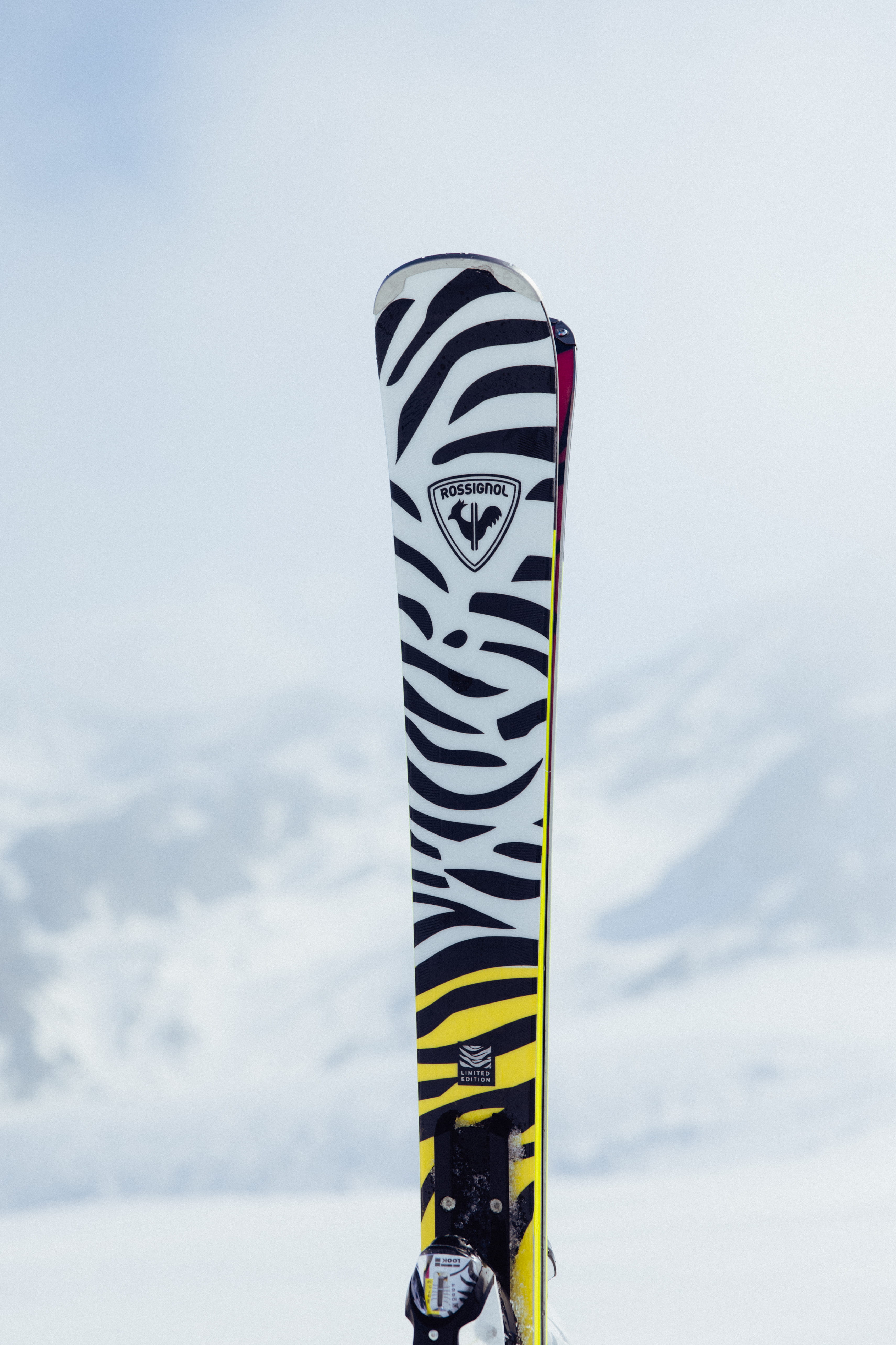 Close-up of a ski with a bold black-and-white graphic standing upright in the snow, with snow-covered mountains in the background.