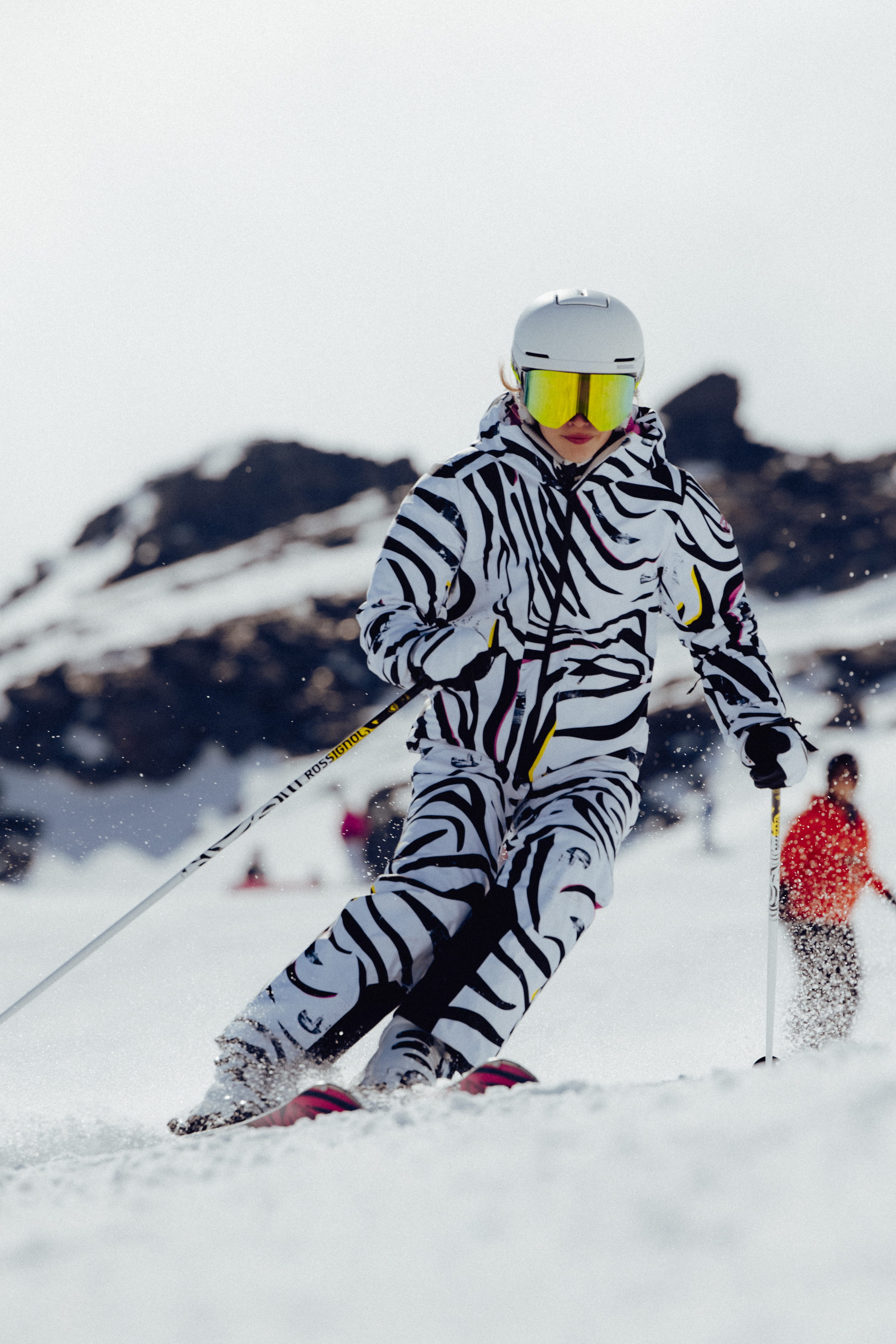 Action shot of a skier in a bold black-and-white patterned ski outfit skiing down a snowy slope, with snow spraying from the skis and mountain peaks in the background.