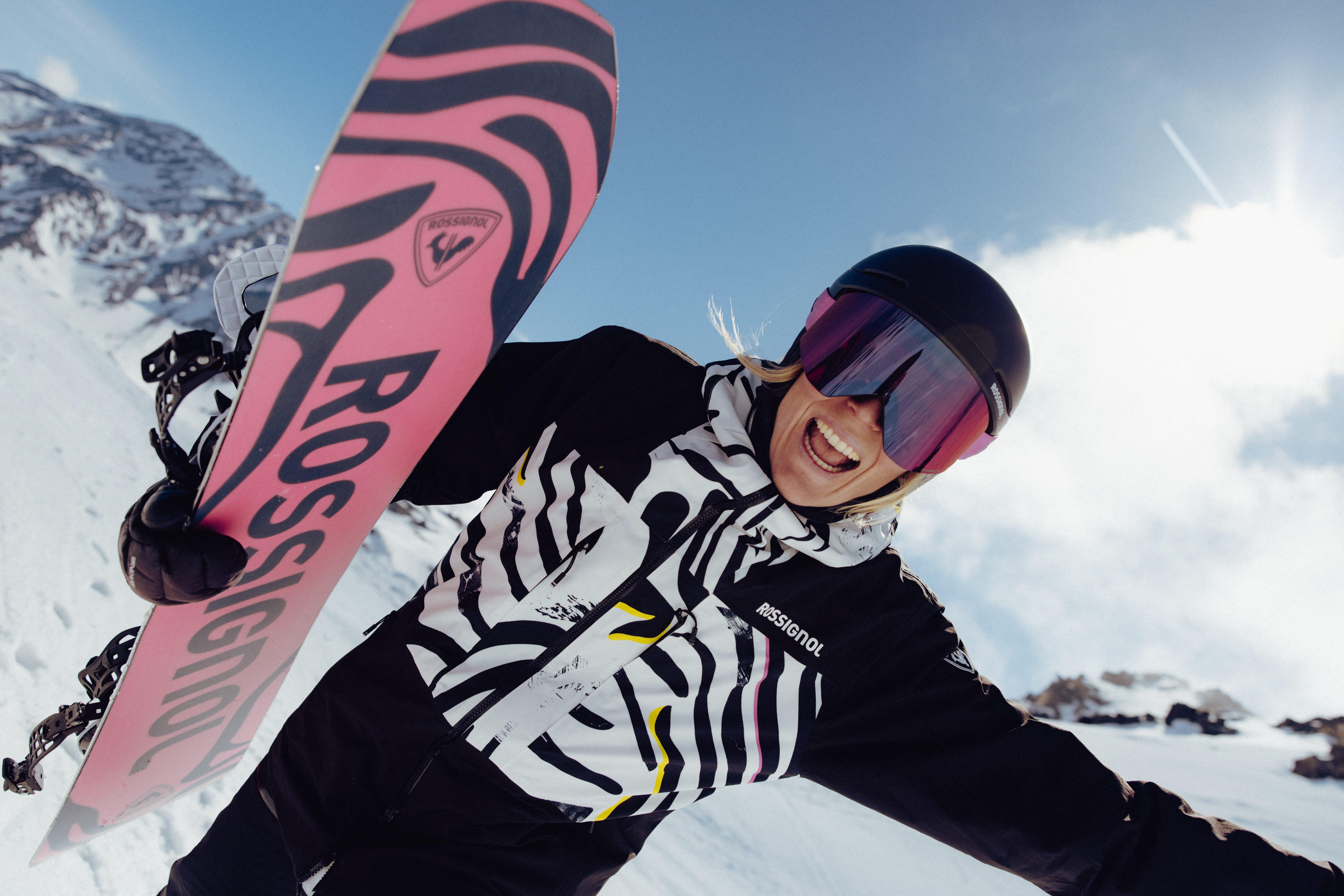 Gros plan d’une personne tenant un snowboard avec un graphisme Rossignol rose vif, portant une veste de neige à motifs noirs et blancs, un casque et un masque à écran miroir sur une pente enneigée en montagne.