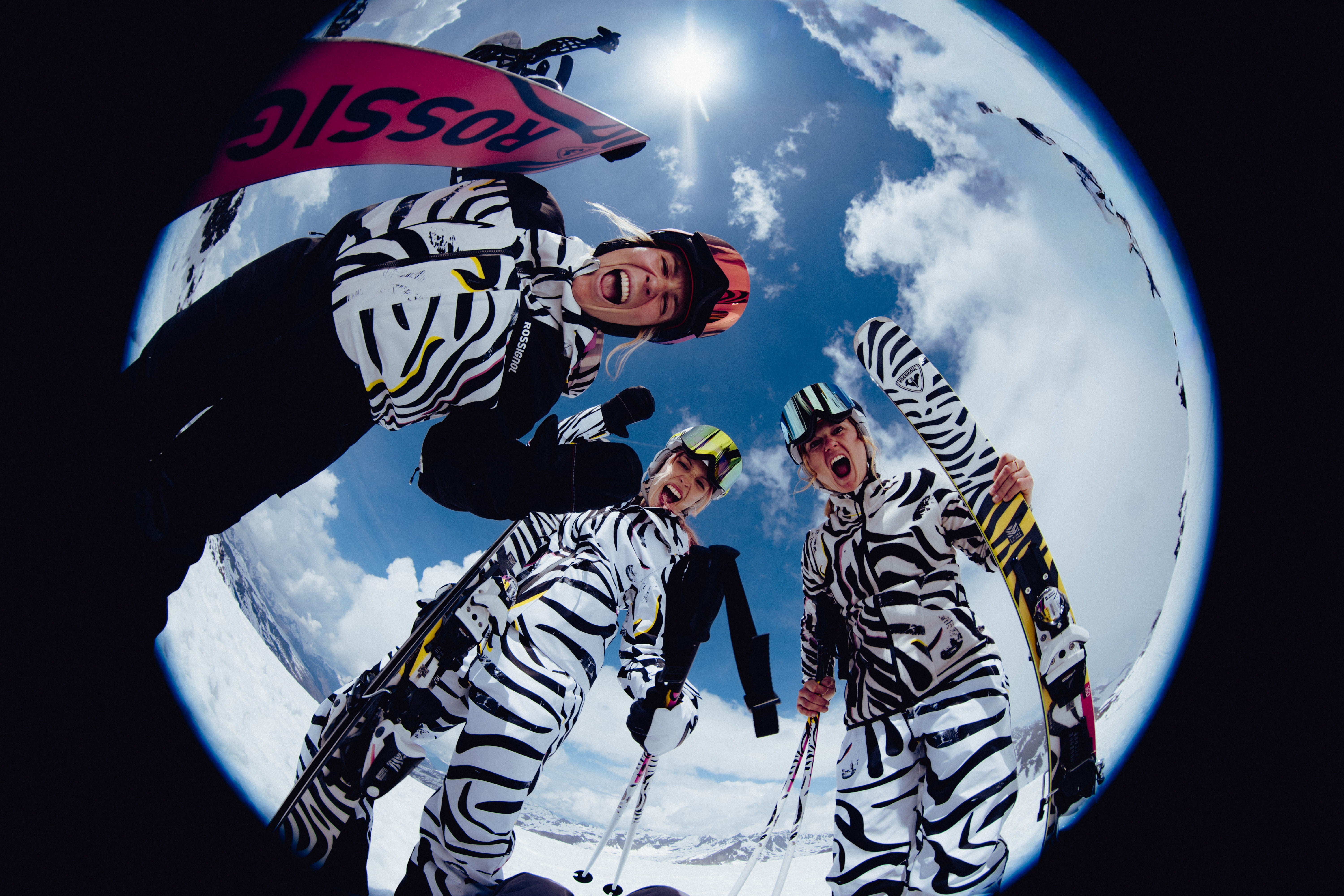 Fisheye view of three skiers in black-and-white patterned ski outfits standing on a snowy mountain slope beneath a bright blue sky.