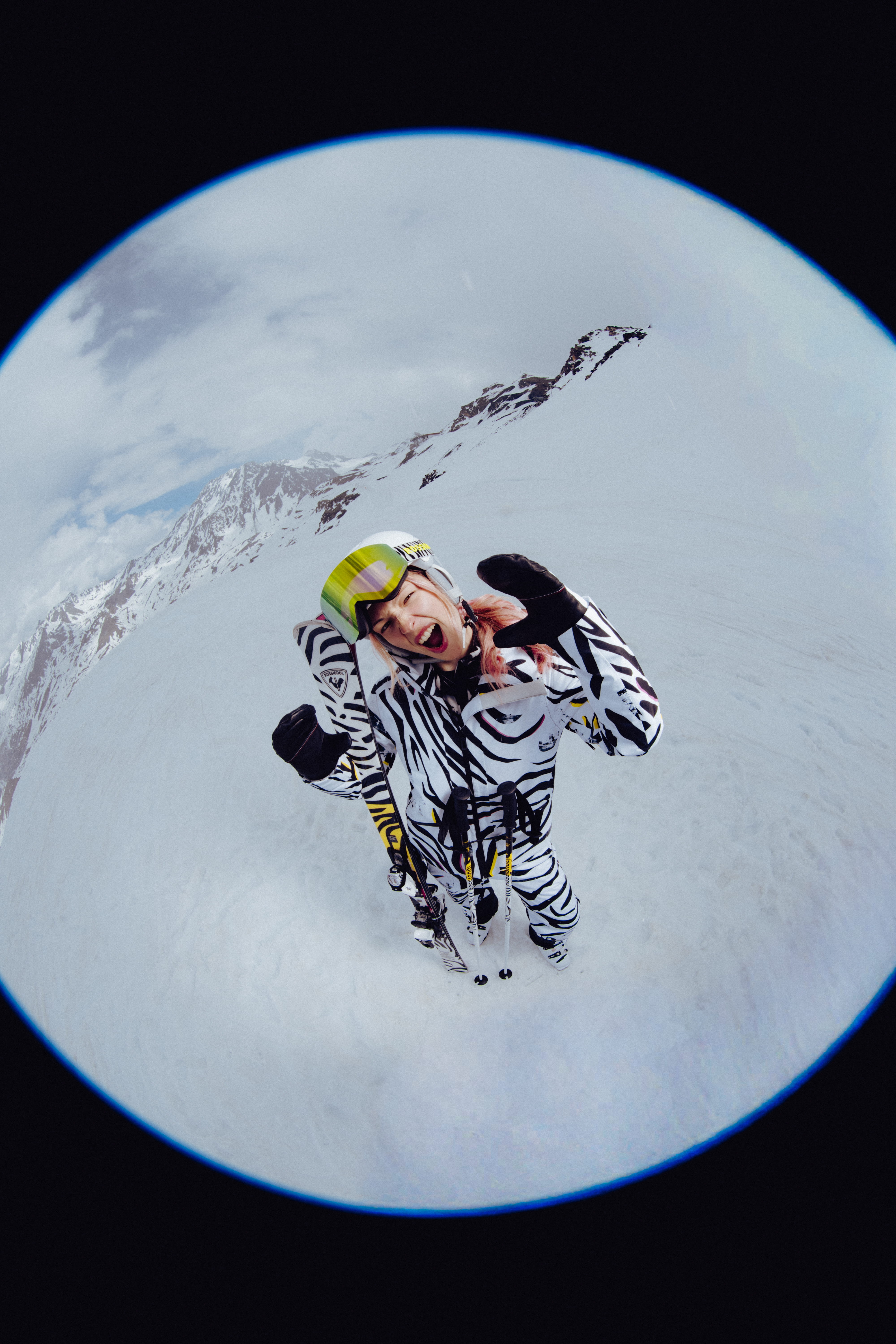 Fisheye view of a skier in a bold black-and-white patterned ski outfit standing on a snowy mountain slope, wearing a white helmet and mirrored goggles.