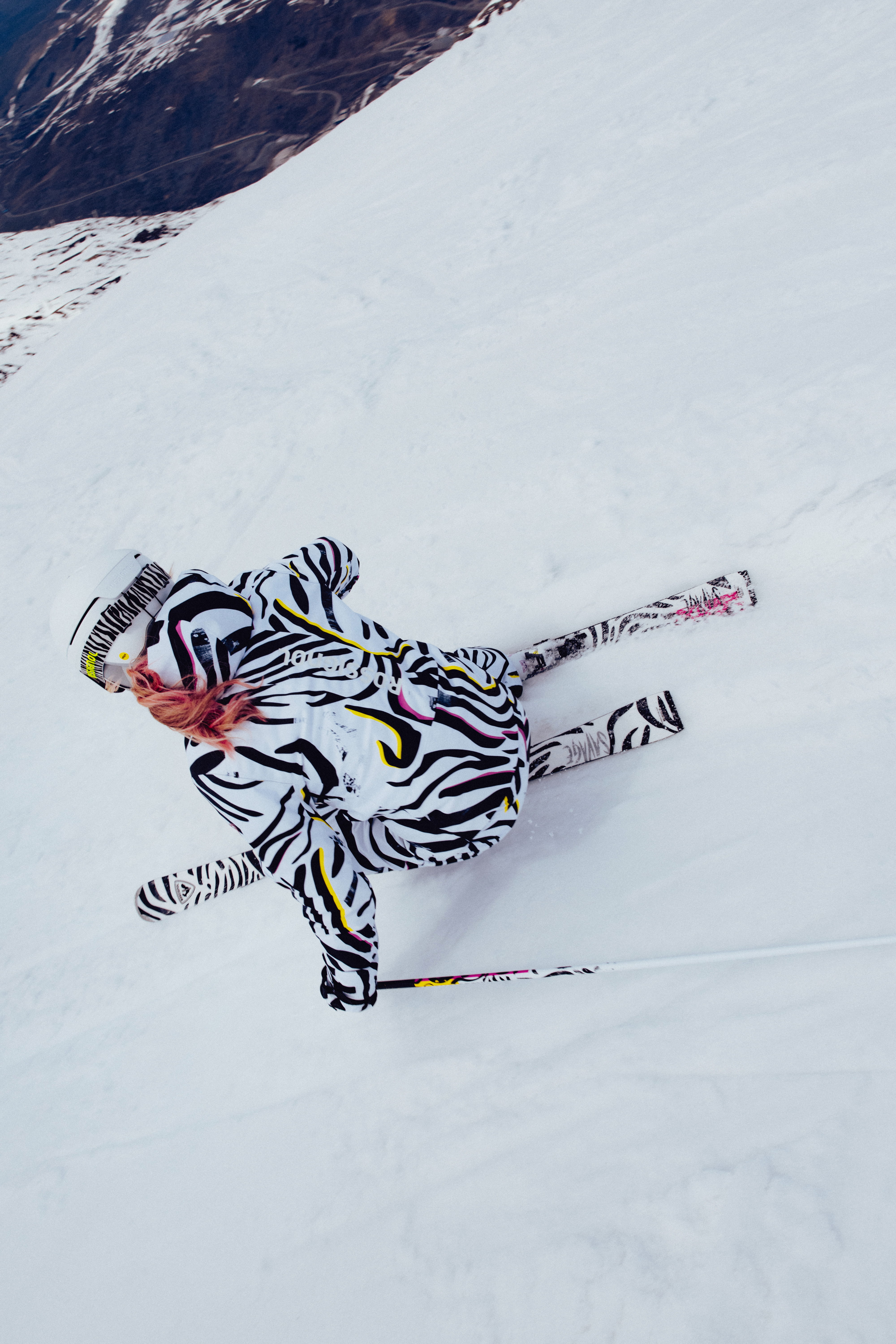 Skier in a bold black-and-white patterned ski outfit descending a mountain slope, viewed from above.