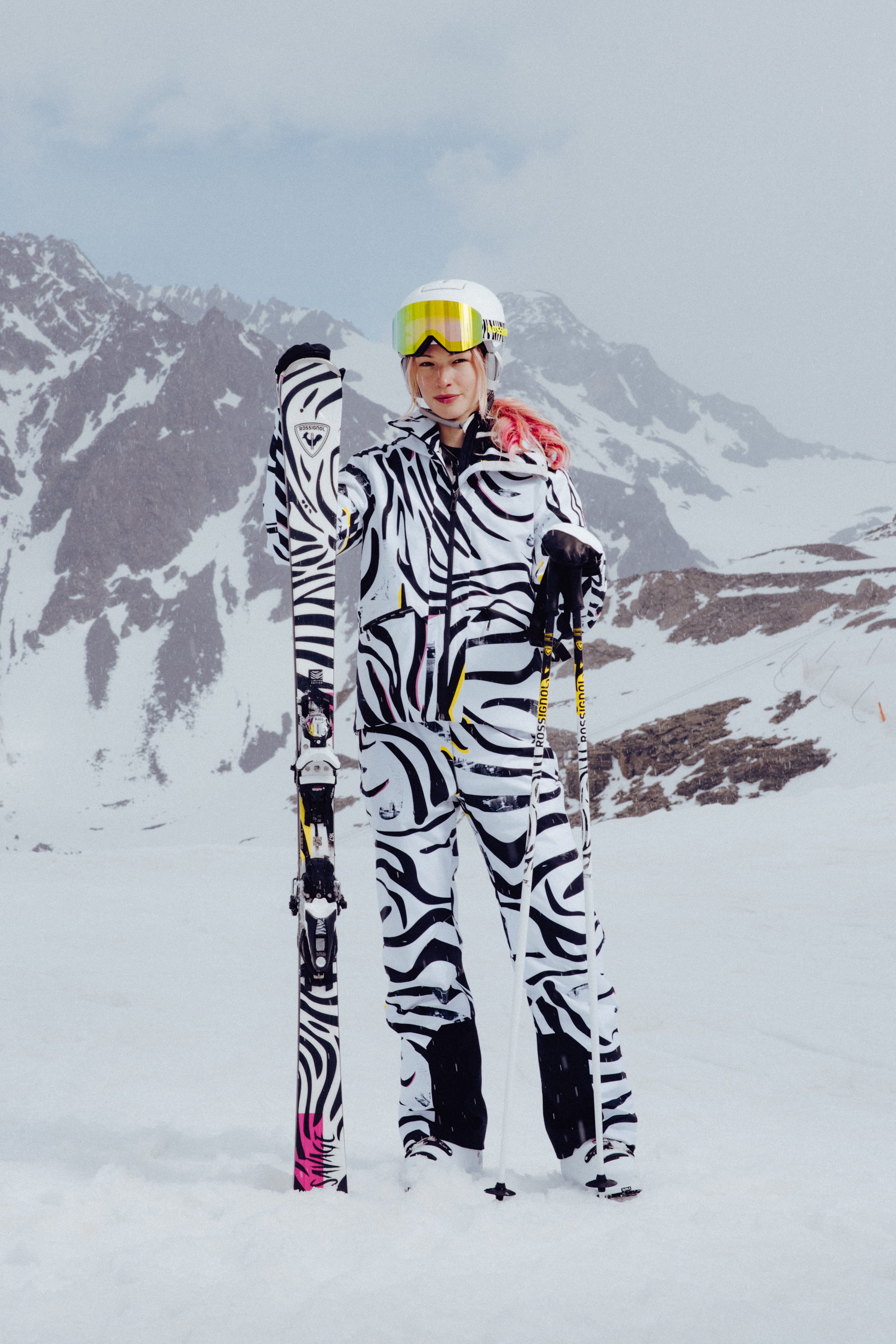 Person standing on a snowy mountain slope wearing a bold black-and-white patterned ski outfit, holding skis and ski poles, with snow-covered peaks in the background.