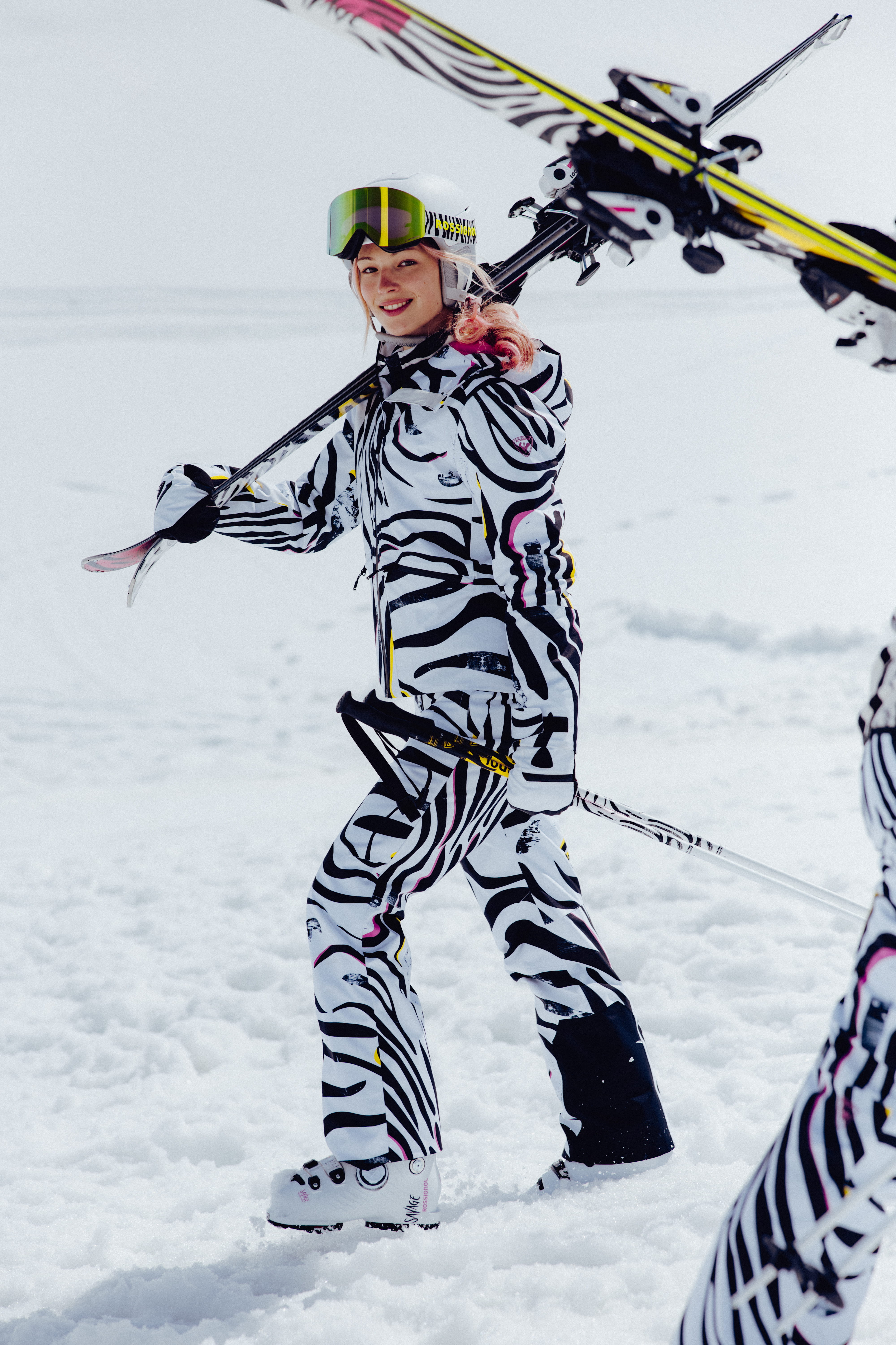 Person carrying skis over a shoulder while walking through the snow, wearing a bold black-and-white patterned ski outfit.