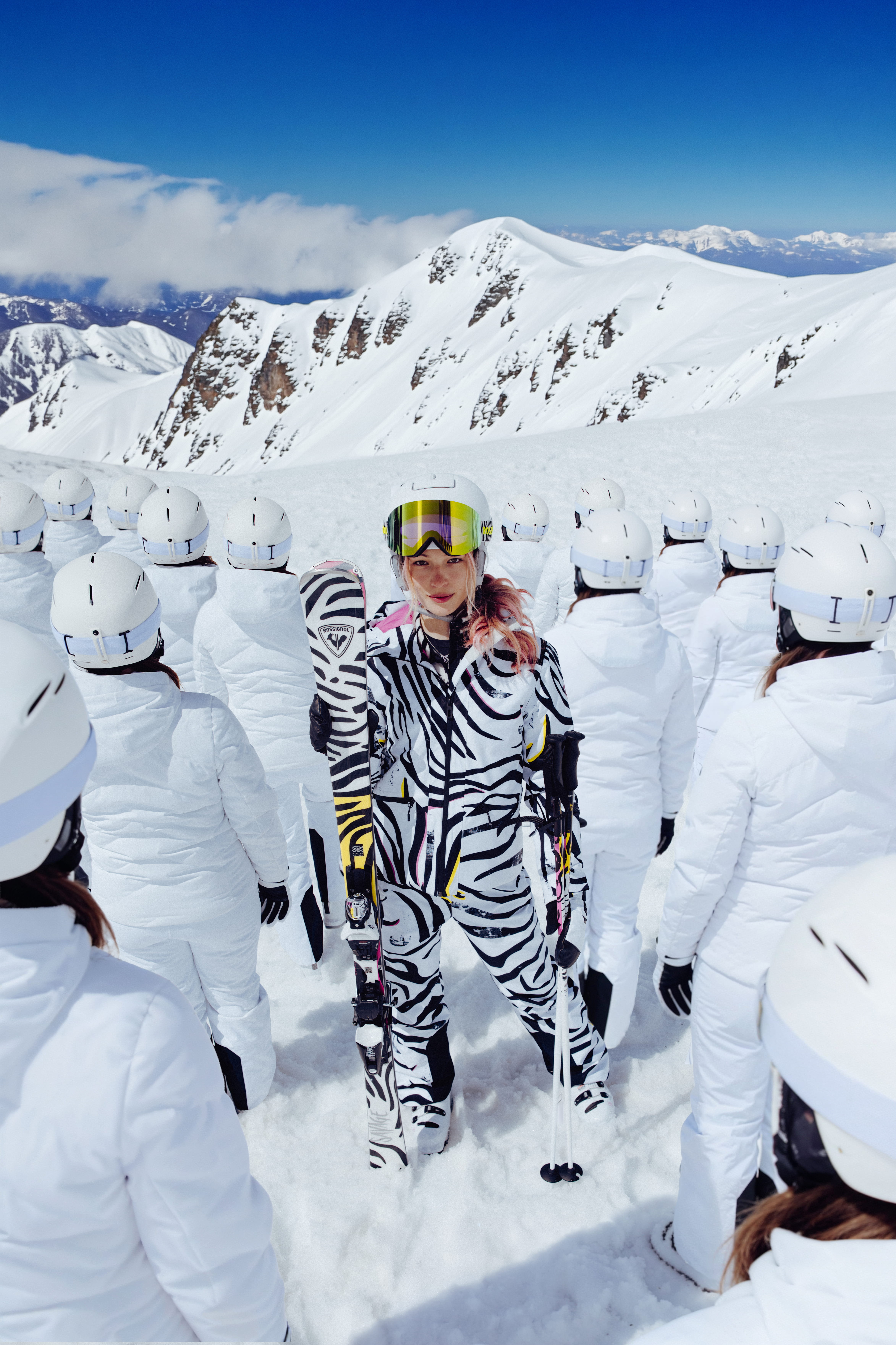 Person wearing a black-and-white patterned ski outfit stands on the snow holding skis, surrounded by people in white ski gear, with snow-covered mountain peaks in the background.